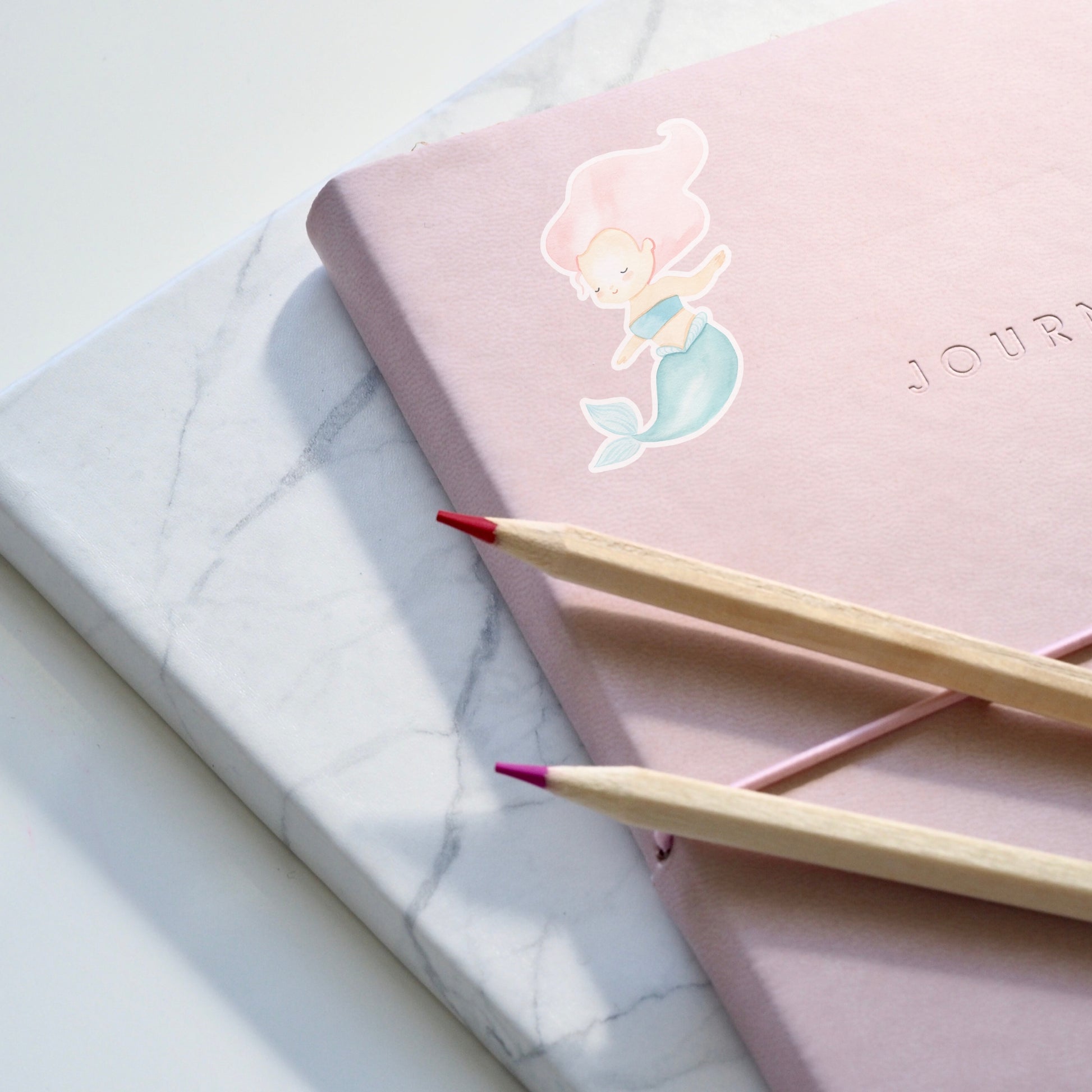 Sticker Meerjungfrau pink: Ein handgemalter, spritzfester Vinyl-Sticker in Form einer Meerjungfrau auf einem pinken Notizbuch, begleitet von zwei Bleistiften.