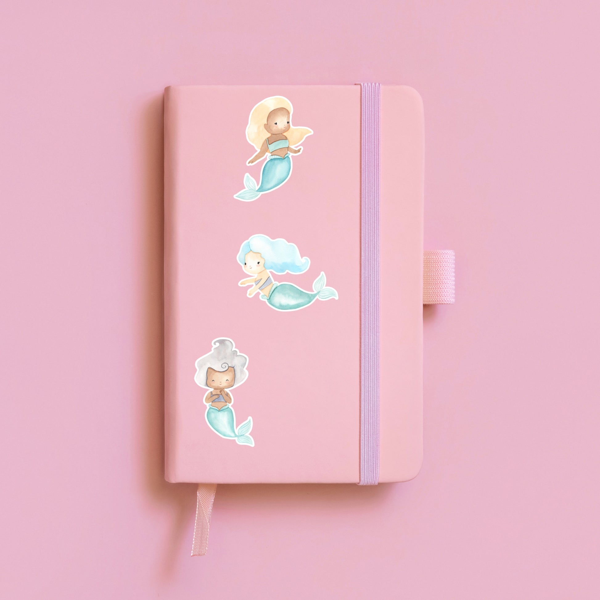 Sticker Meerjungfrau blond auf rosa Notizbuch mit verschiedenen Stickern, zeigt handgemalte Meerjungfrau in Pastelltönen, ideal für Bullet Journals oder Schulhefte.