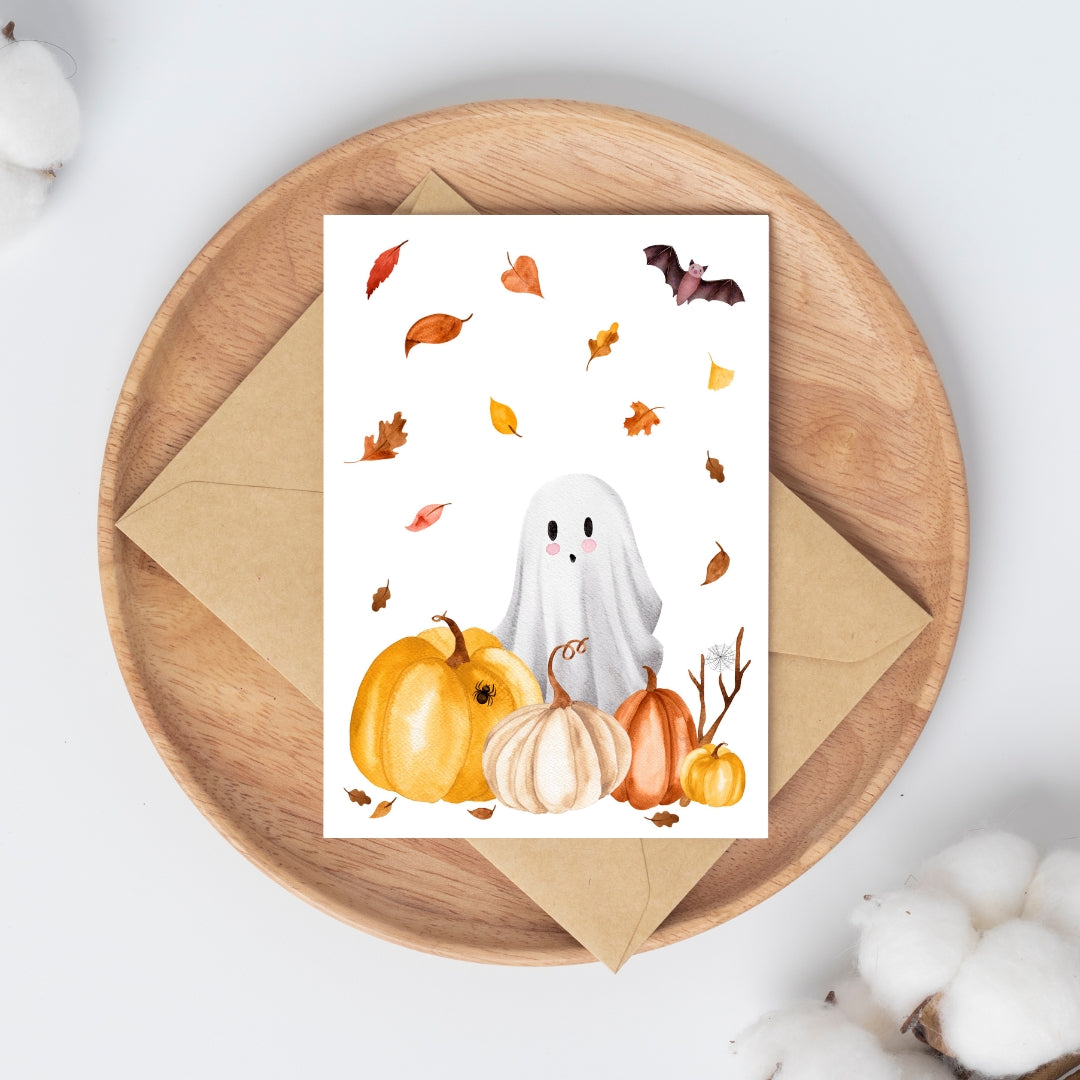 Handgemalte Halloweenkarte mit Aquarell-Gespenst, Kürbissen, Herbstlaub und Fledermaus auf braunem Umschlag auf Holzbrett.