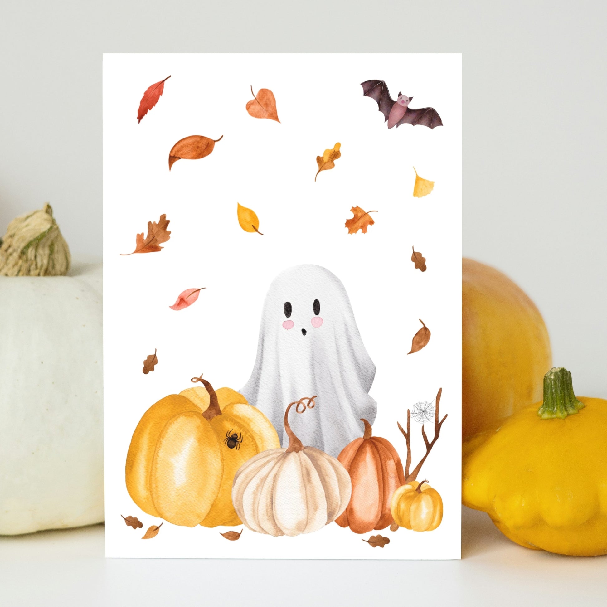 Handgemalte Halloweenkarte mit Aquarell-Gespenst, Kürbissen, Herbstlaub und Fledermaus an echte Kürbisse gelehnt.
