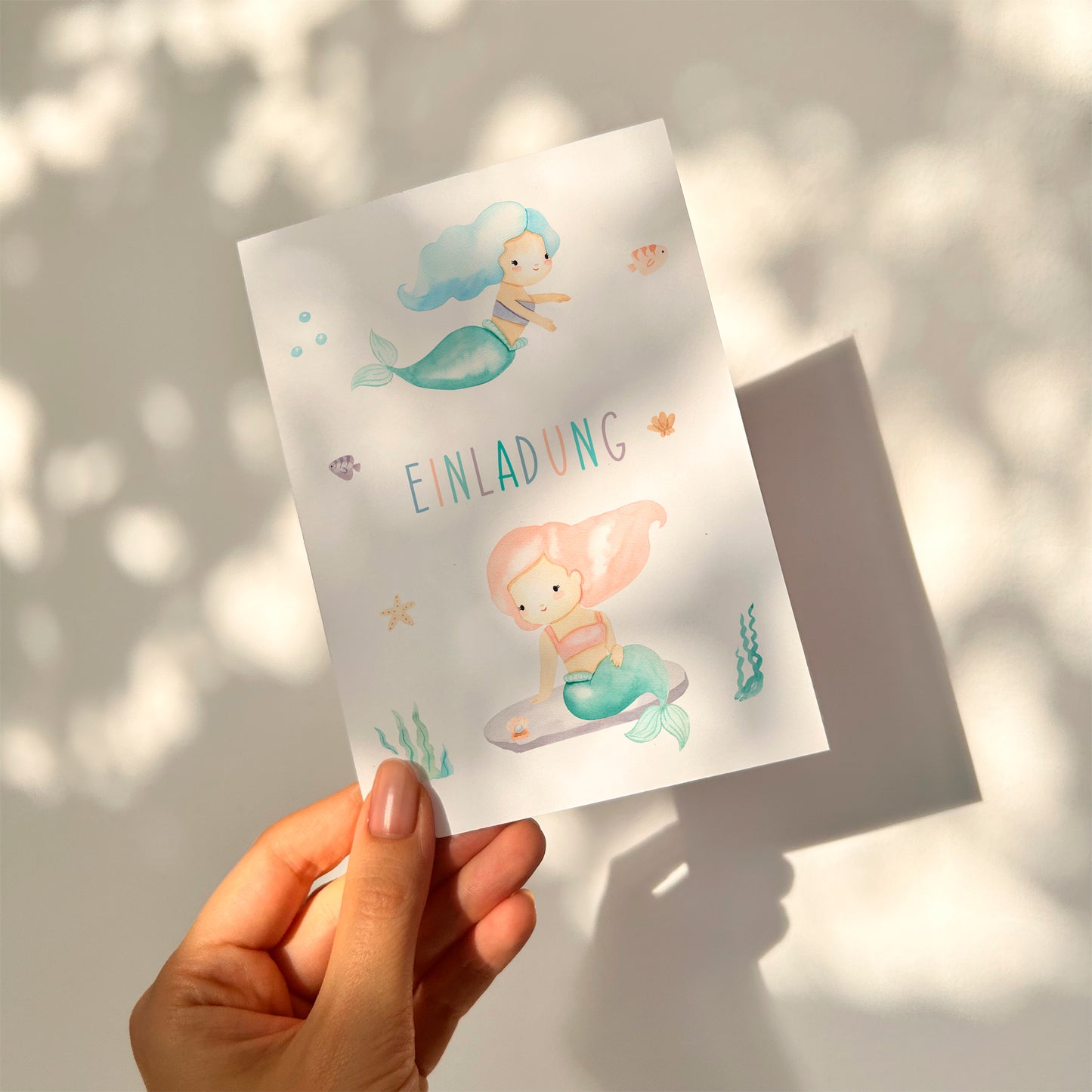 Einladung Kindergeburtstag Meerjungfrauen A6: Hand hält pastellfarbene Karte mit zwei Aquarell-Meerjungfrauen und maritimen Motiven, EINLADUNG in der Mitte.