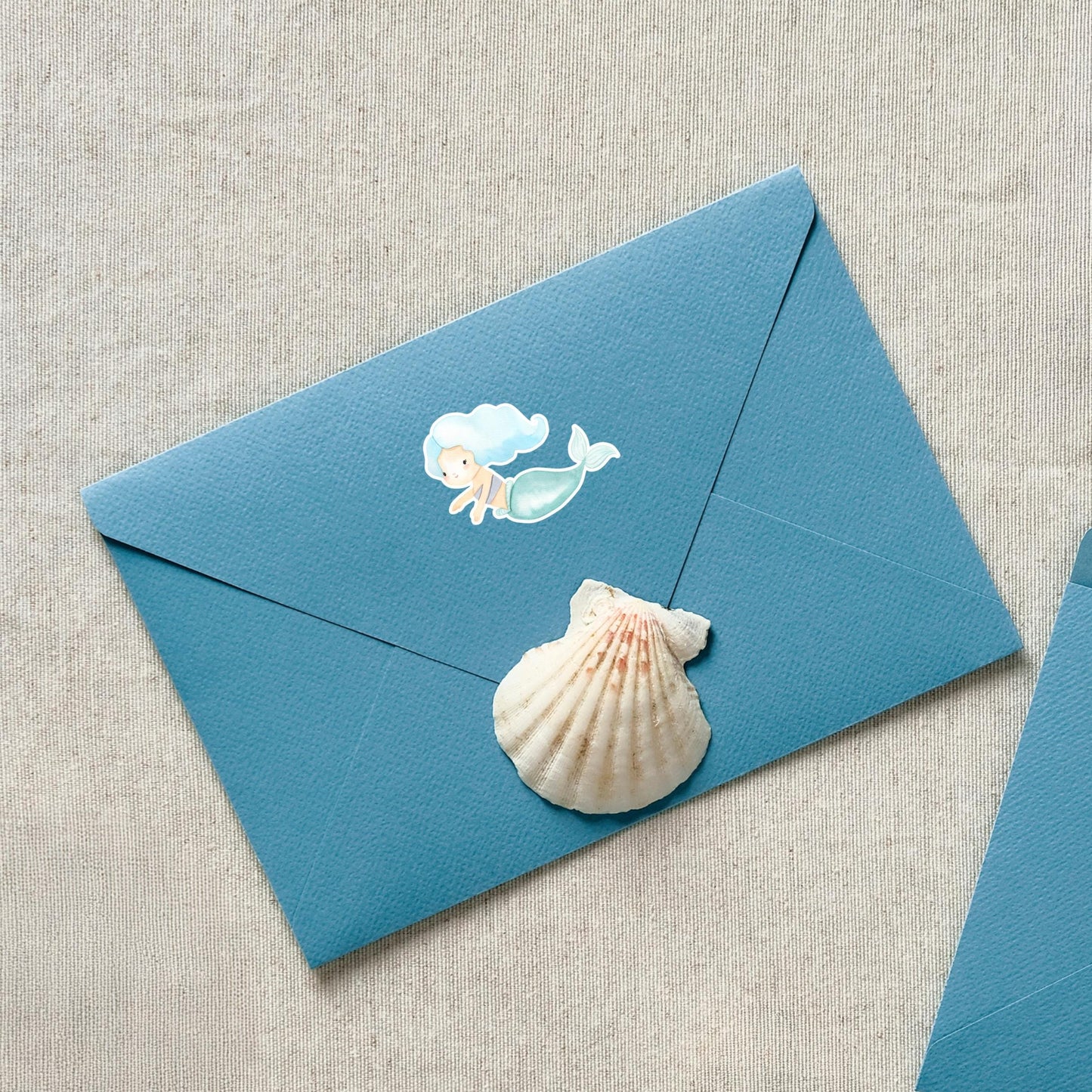 Sticker Meerjungfrau blau auf blauem Briefumschlag mit Muschel, zeigt handgemalten Meerjungfrau-Sticker in Pastelltönen. Ideal zur Dekoration von Notizbüchern und Laptops.