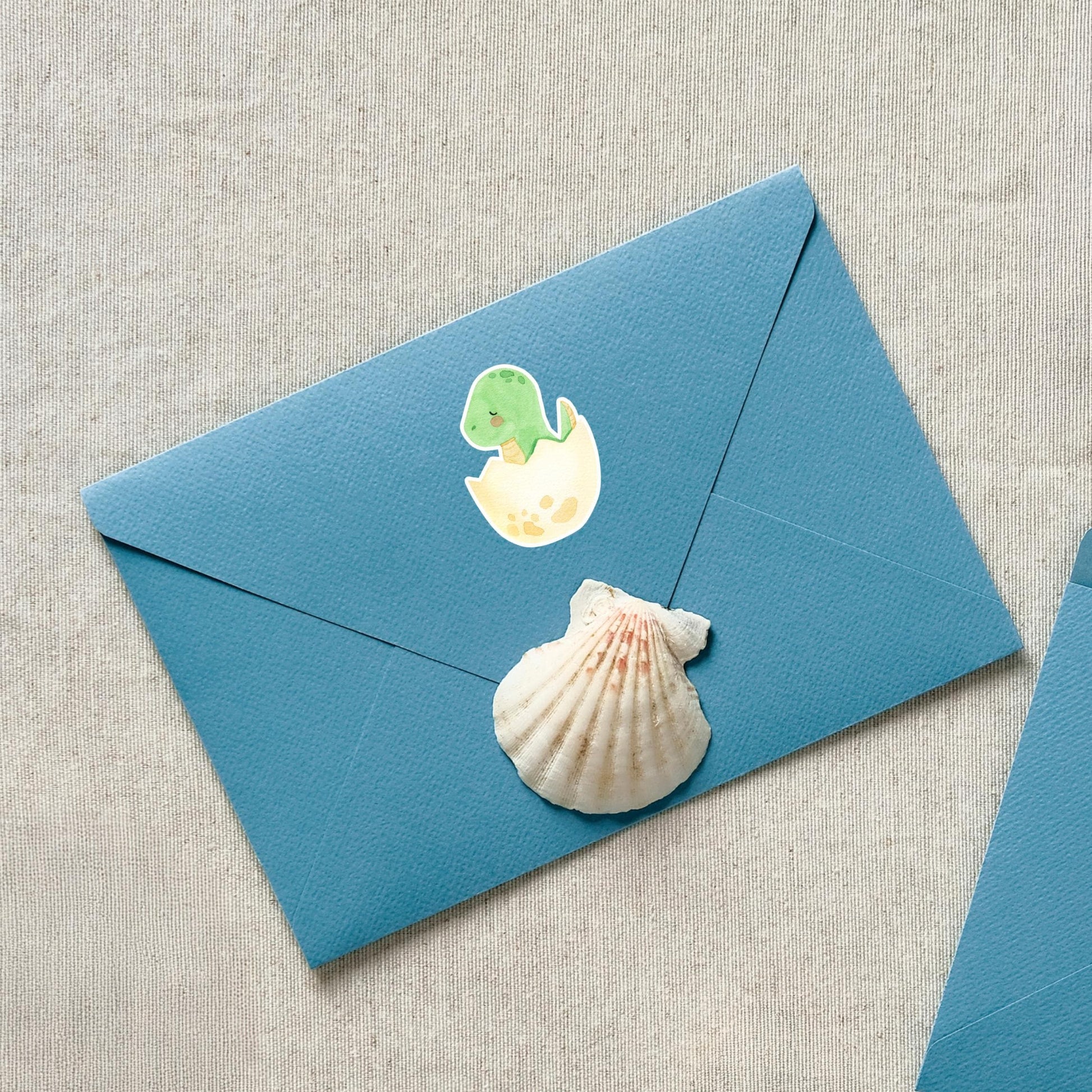 Sticker Dinosaurier im Ei: Ein grüner Dinosaurier schlüpft aus einem Ei auf einem blauen Umschlag mit Muschelmotiv. Hochwertig und ideal für Bastelprojekte.