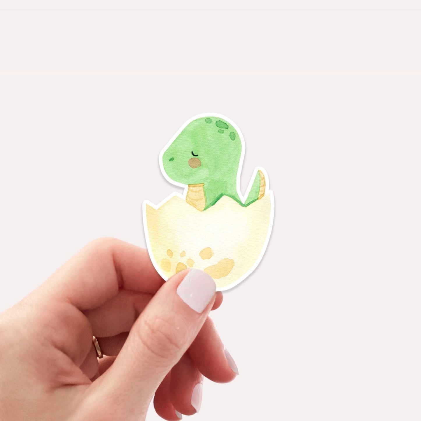 Sticker Dinosaurier im Ei: Eine Hand hält einen handgemalten Vinyl-Sticker mit einem grünen Dinosaurier, der aus einem Ei schlüpft. Perfekt für individuelle Dekoration.