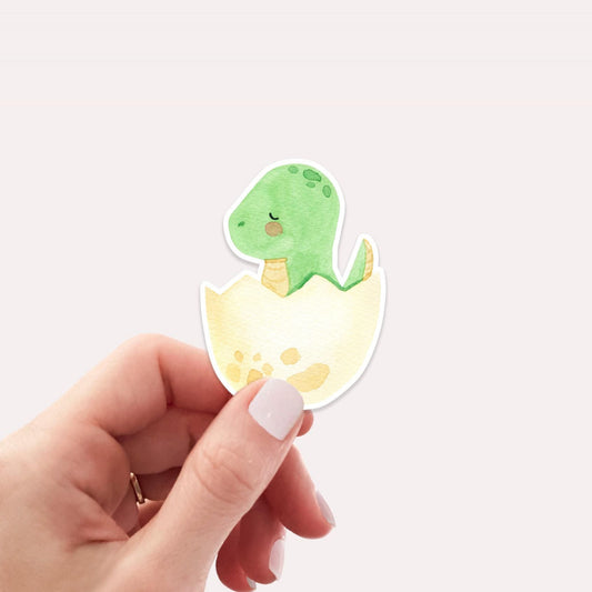 Sticker Dinosaurier im Ei: Eine Hand hält einen handgemalten Vinyl-Sticker mit einem grünen Dinosaurier, der aus einem Ei schlüpft. Perfekt für individuelle Dekoration.