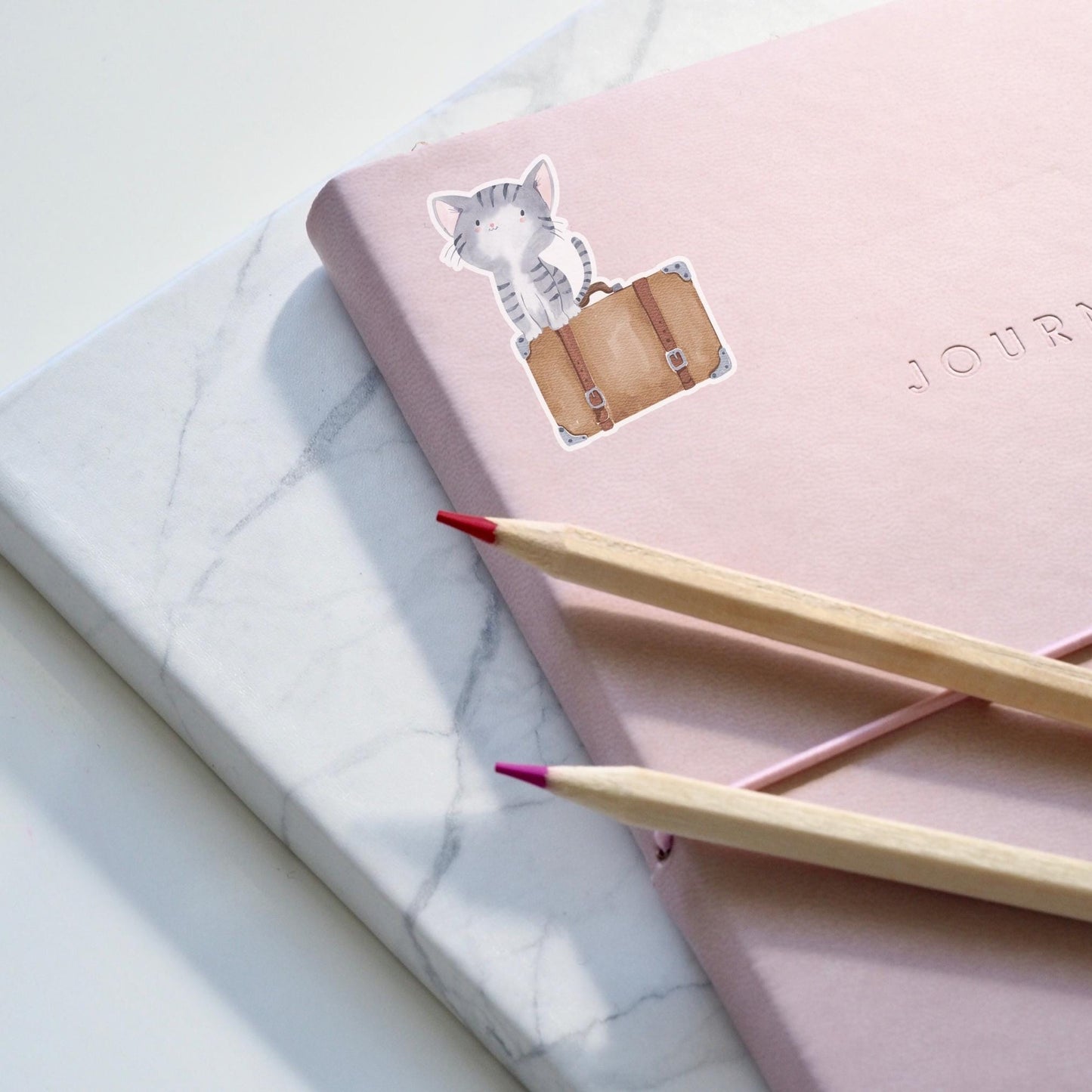 Sticker Katze & Koffer: Ein handgezeichneter Katzen-Vinyl-Sticker neben einem pinken Notizbuch, ideal zum Dekorieren von Journalen und Accessoires.