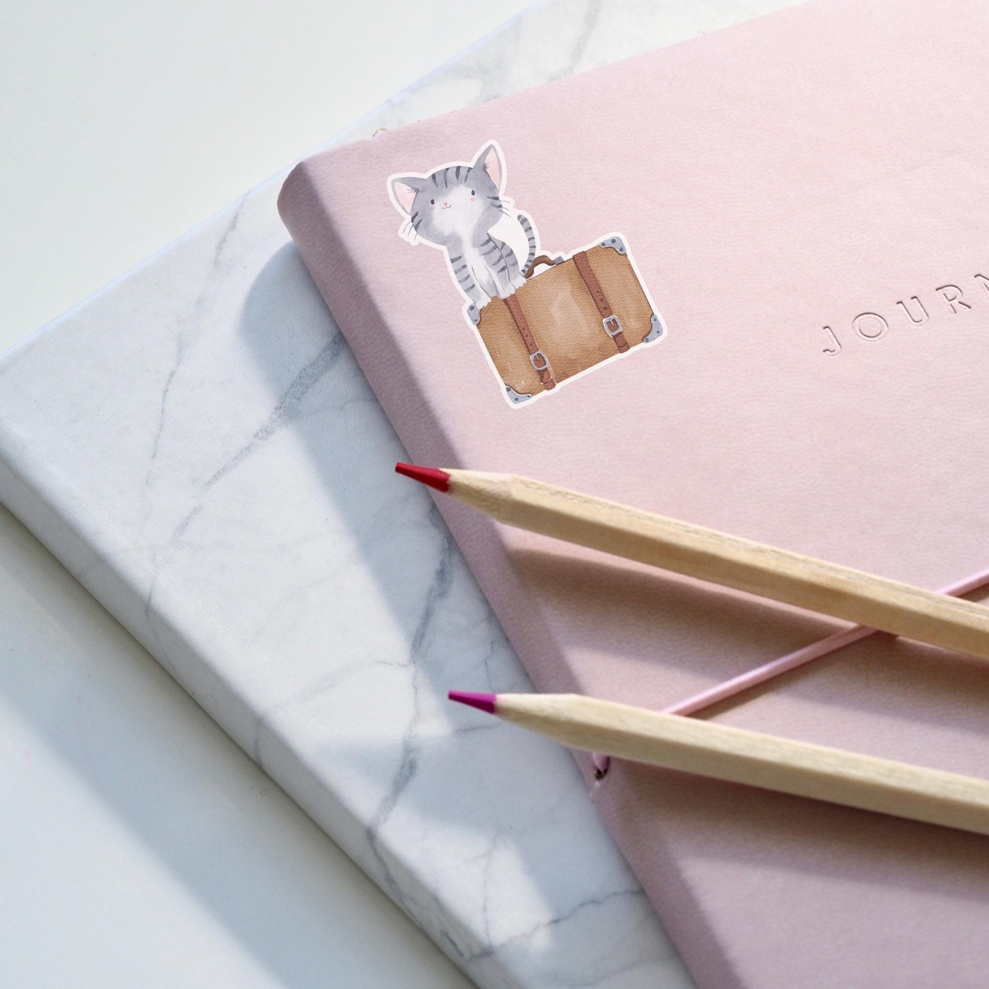 Sticker Katze & Koffer: Ein handgezeichneter Katzen-Vinyl-Sticker neben einem pinken Notizbuch, ideal zum Dekorieren von Journalen und Accessoires.
