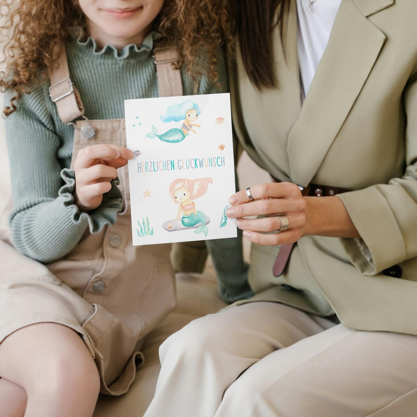 Frau hält eine Karte Kindergeburtstag Meerjungfrauen mit handgemalten Meerjungfrauen und Herzlichen Glückwunsch Schriftzug, ideal für Kindergeburtstage.