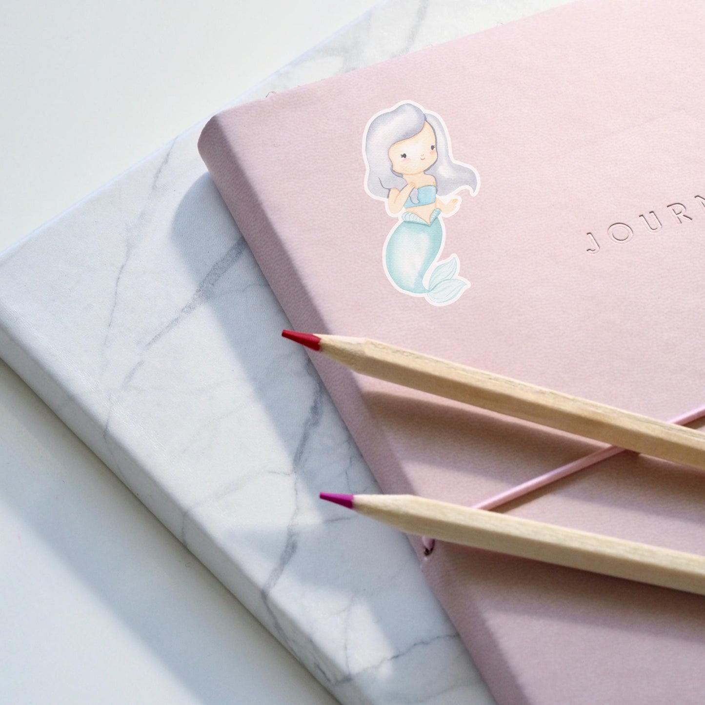 Sticker Meerjungfrau grau auf einem pinken Notizbuch, begleitet von Bleistiften. Hochwertiger, langlebiger Vinyl-Sticker, ideal für kreative Dekorationen und persönliche Accessoires.