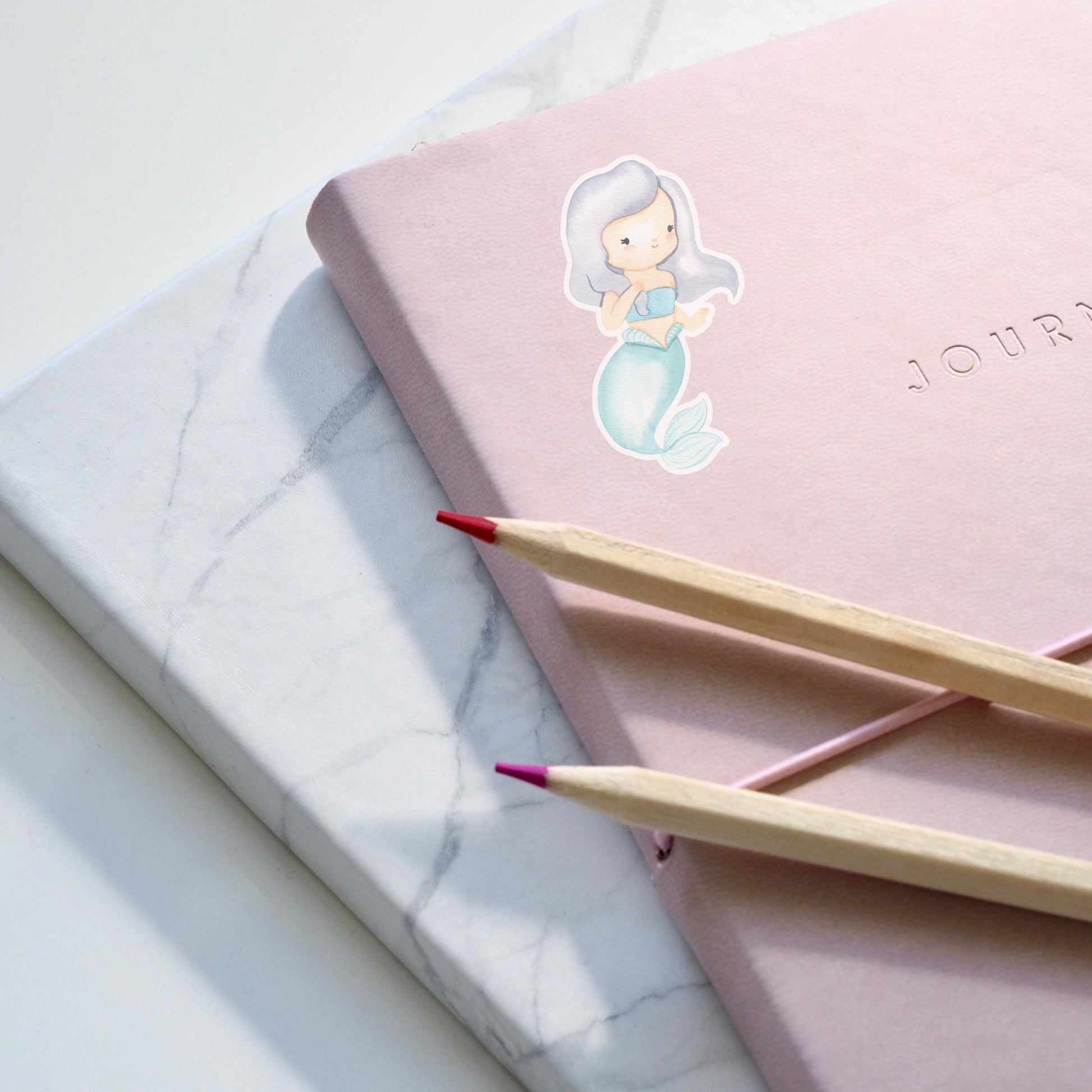 Sticker Meerjungfrau grau auf einem pinken Notizbuch, begleitet von Bleistiften. Hochwertiger, langlebiger Vinyl-Sticker, ideal für kreative Dekorationen und persönliche Accessoires.
