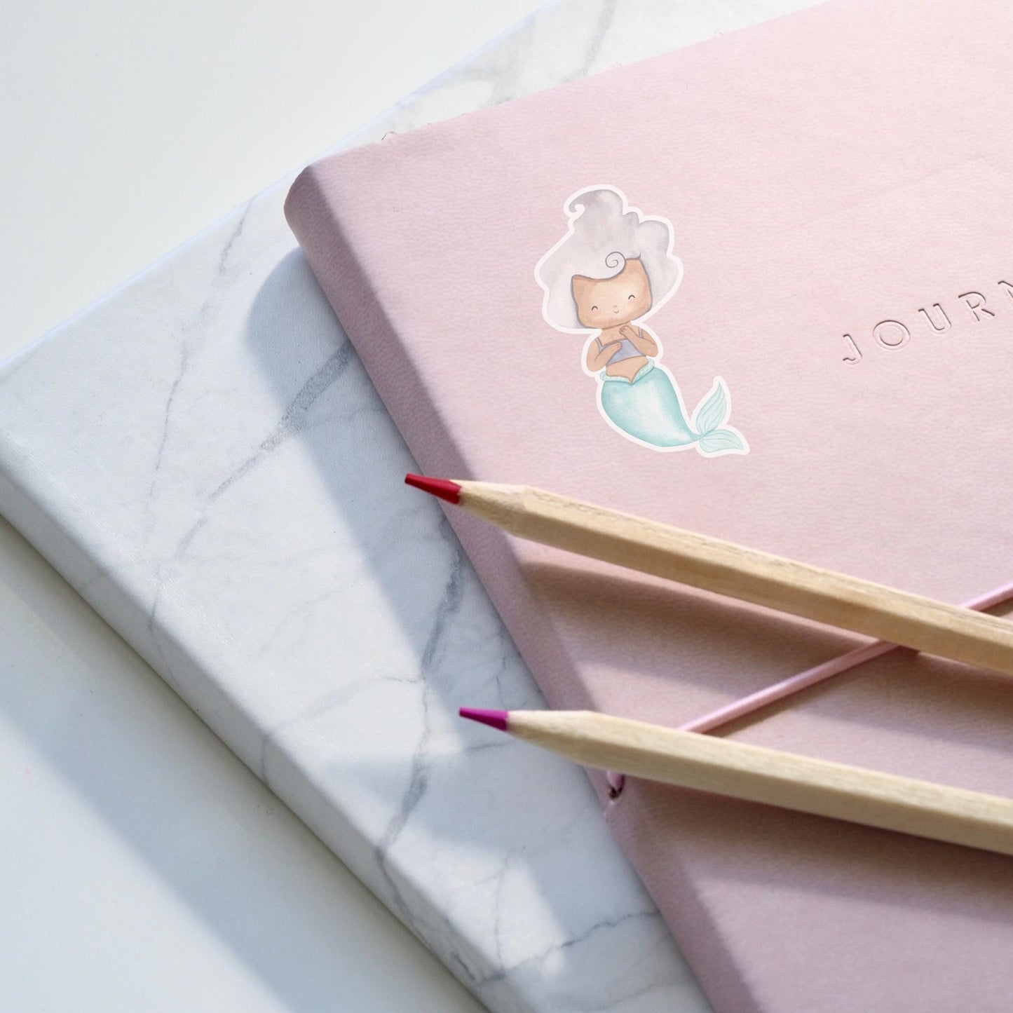 Sticker Meerjungfrau silber auf einem pinken Notizbuch mit zwei Bleistiften, zeigt handgemaltes Meerjungfrauen-Design in Pastell, ideal für kreative Dekorationen.