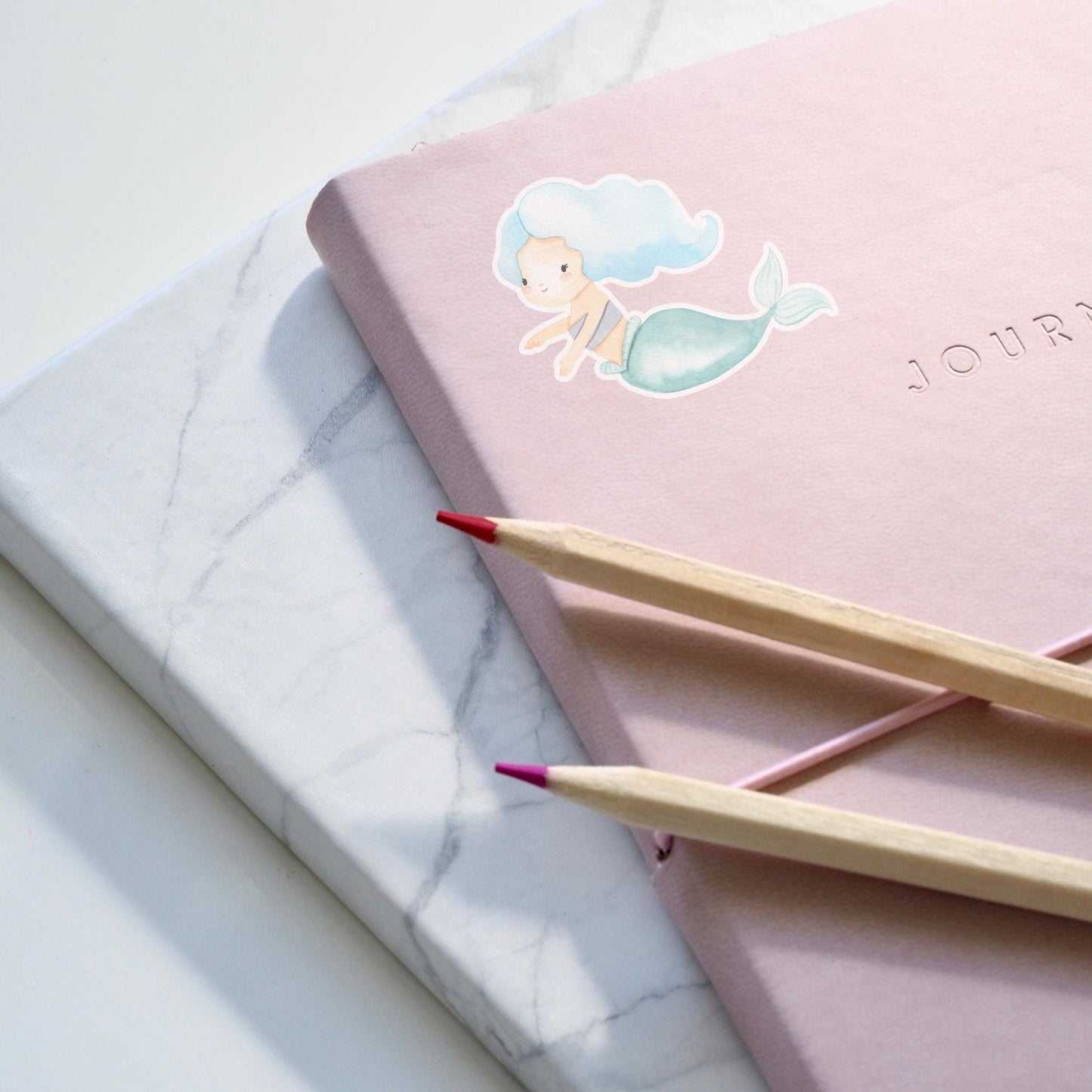 Sticker Meerjungfrau blau: Pinkes Notizbuch mit mermaid Motiv und zwei Bleistiften, zeigt handgemalten, langlebigen Vinyl-Sticker, ideal für kreative Anwendungen und Dekorationen.