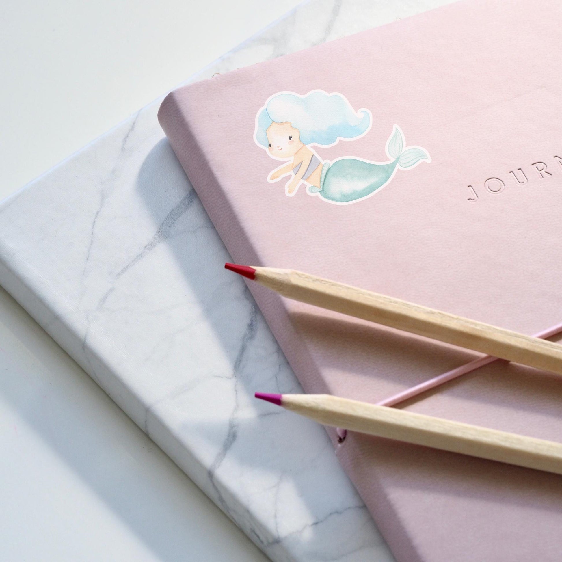 Sticker Meerjungfrau blau: Pinkes Notizbuch mit mermaid Motiv und zwei Bleistiften, zeigt handgemalten, langlebigen Vinyl-Sticker, ideal für kreative Anwendungen und Dekorationen.