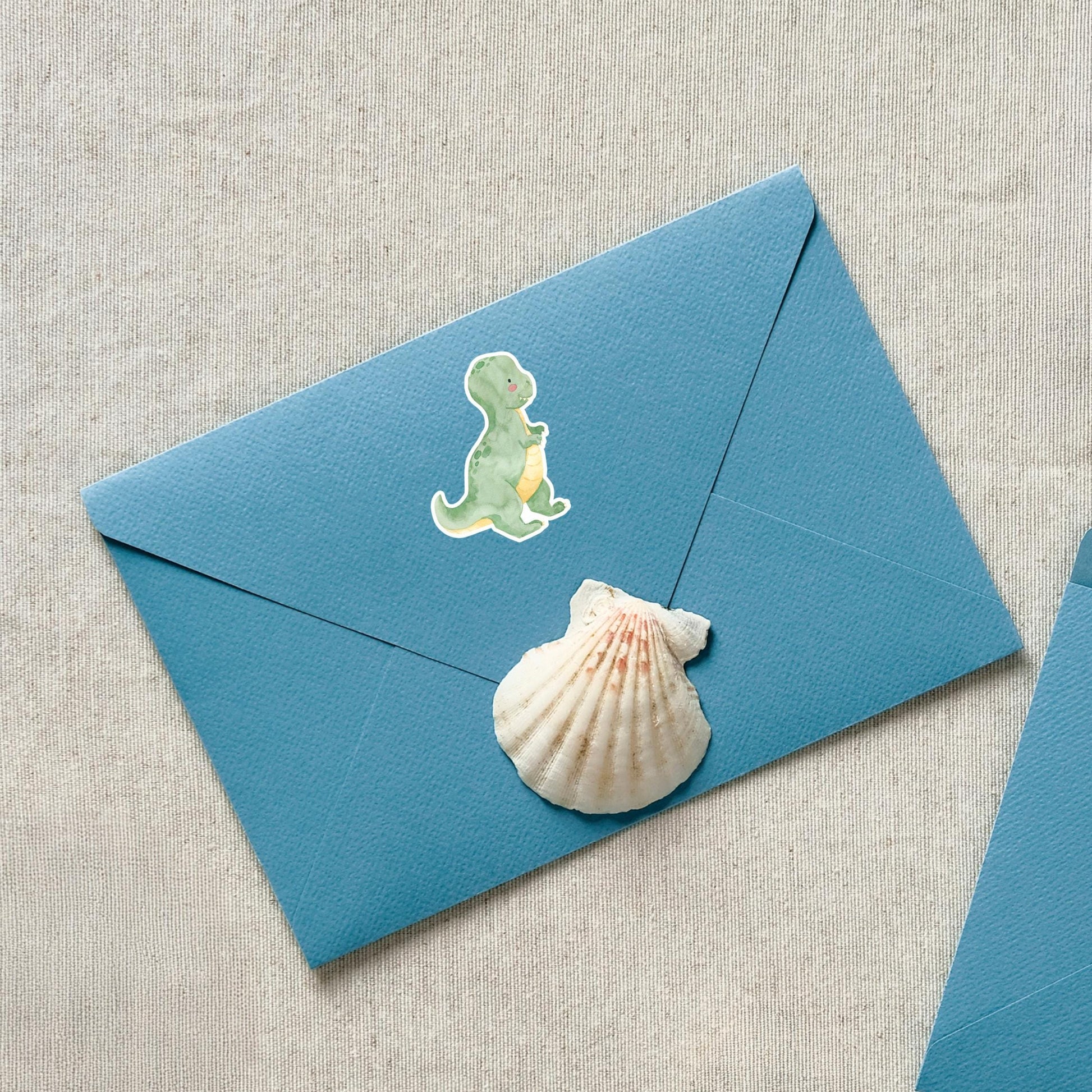 Sticker Dinosaurier Tyranno: Blauer Umschlag mit Dinosaurier- und Muschel-Sticker. Handgezeichnet, spritzfest, ideal für Bullet Journals oder Einladungen. Größe: ca. 6 x 4,5 cm.