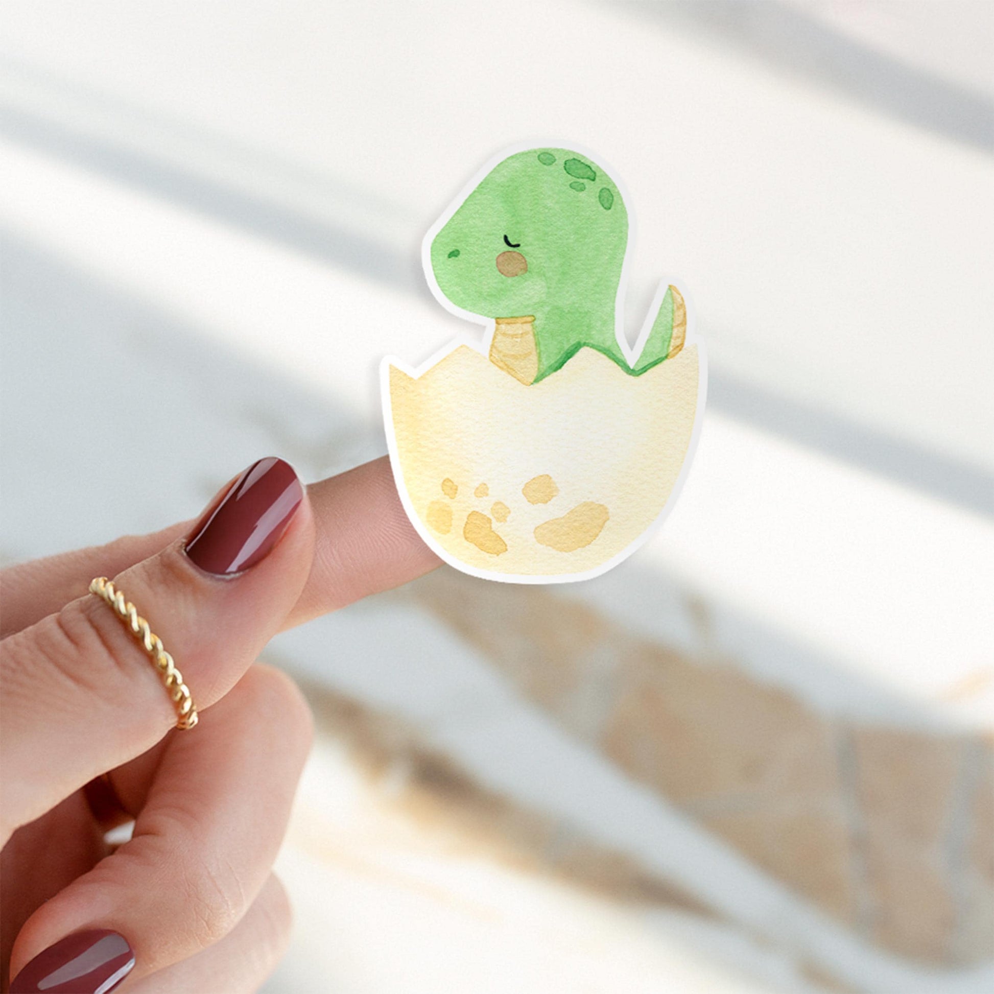 Ein Finger hält den Sticker Dinosaurier im Ei, ein detailreicher, langlebiger Vinyl-Aufkleber ideal für kreative Projekte und individuelle Designs.