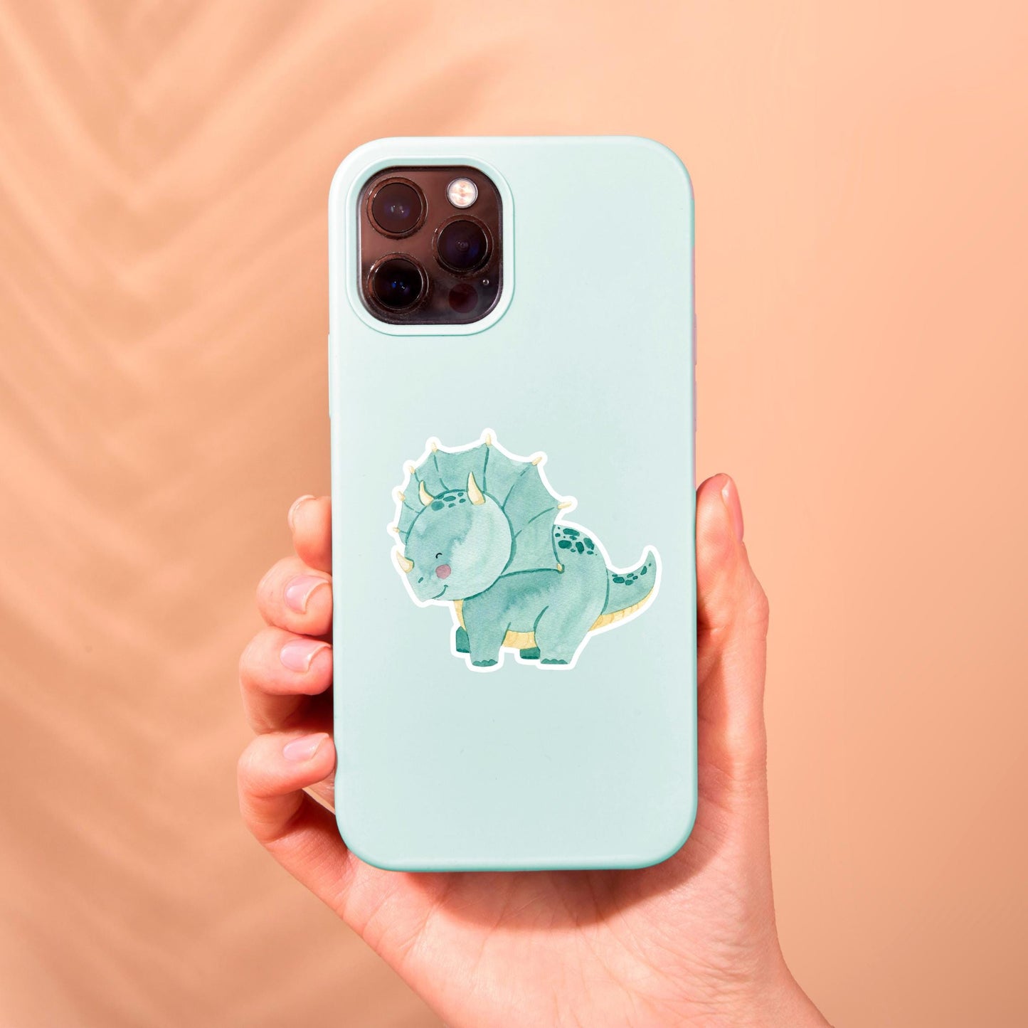 Hand hält eine Handyhülle mit einem Sticker Dinosaurier Dreizack, einem handgemalten, langlebigen Vinyl-Sticker, ideal für individuelle Gestaltung.