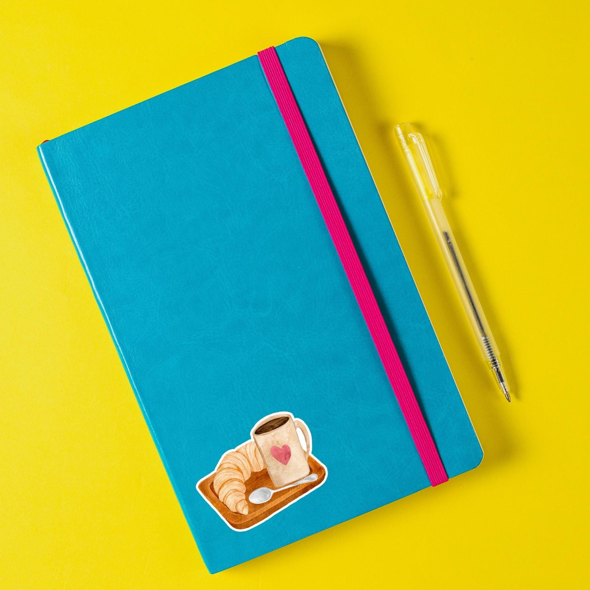 Sticker Frühstück: Ein blauer Notizblock mit rosa Band und Stift, inspiriert von Kaffee und Croissant, ideal zur Dekoration von Bullet Journals und Laptops.