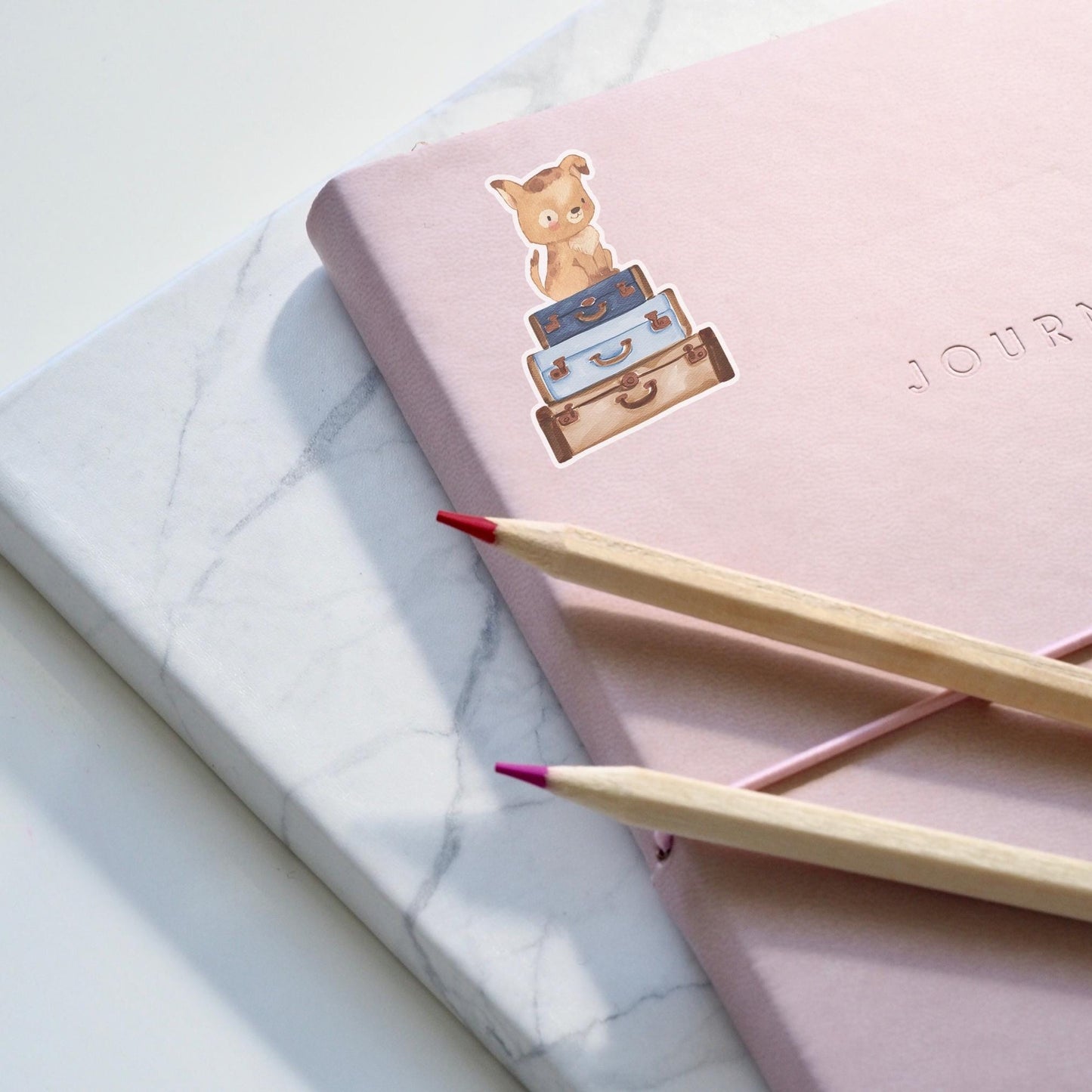 Sticker Hund & Koffer: Handgemalter Hunde-Sticker auf einem rosa Notizbuch mit Bleistiften, inspiriert zu Reisen, ideal für Bullet Journals und DIY-Projekte.
