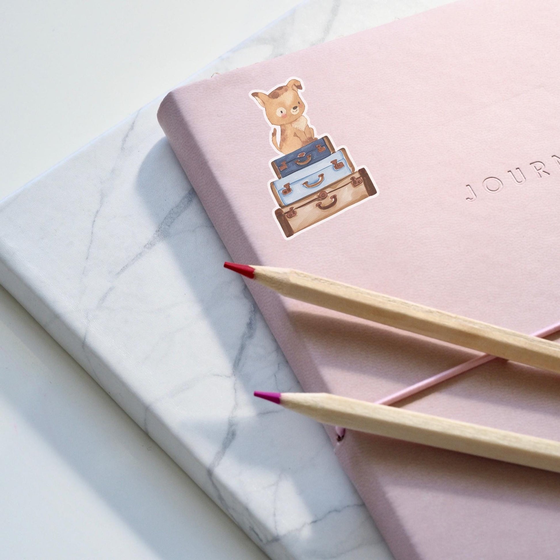 Sticker Hund & Koffer: Handgemalter Hunde-Sticker auf einem rosa Notizbuch mit Bleistiften, inspiriert zu Reisen, ideal für Bullet Journals und DIY-Projekte.