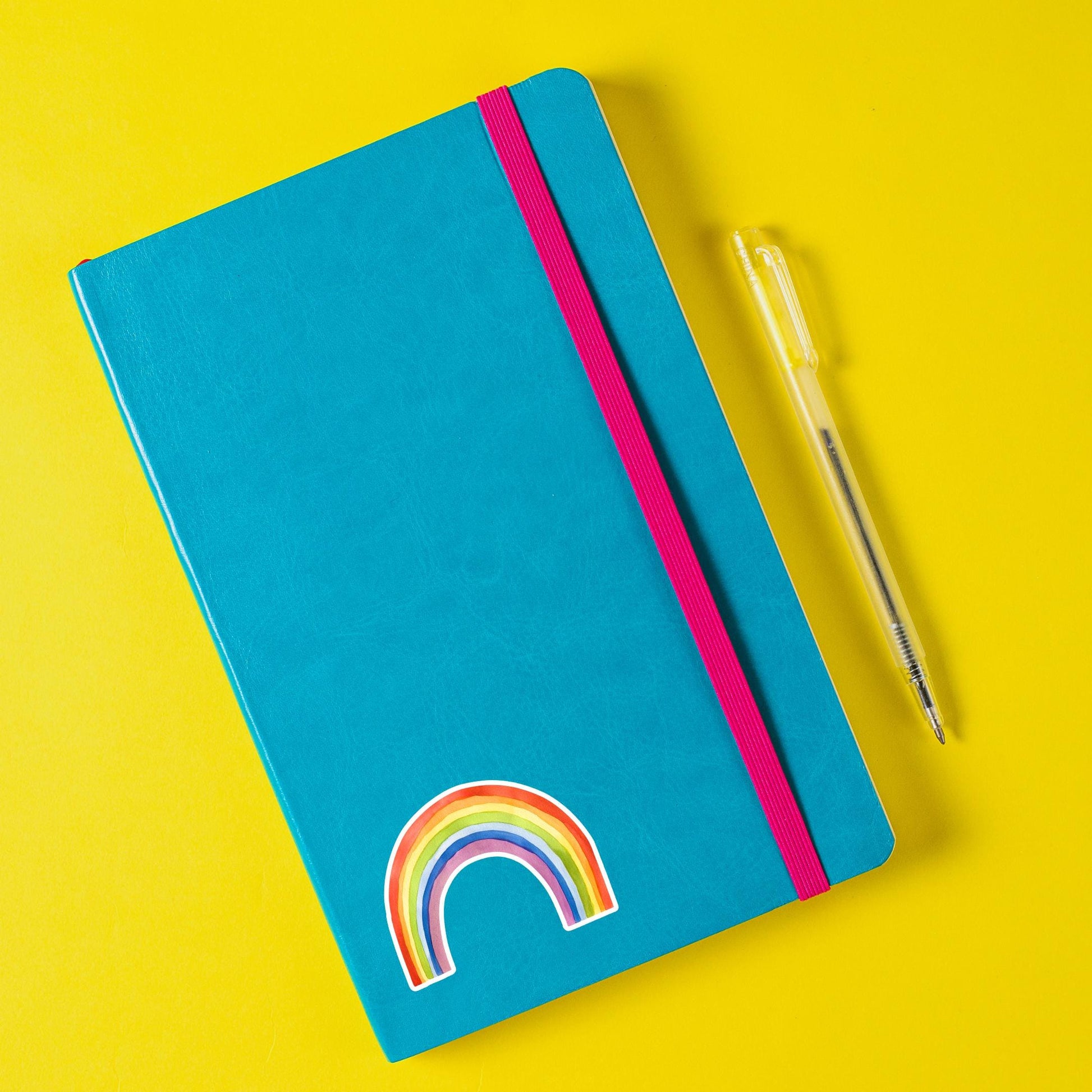 Geschlossener Notizbuch mit Sticker Regenbogen auf dem Cover, daneben ein Kugelschreiber. Der handgemalte Vinyl-Sticker ist spritzfest und langlebig.