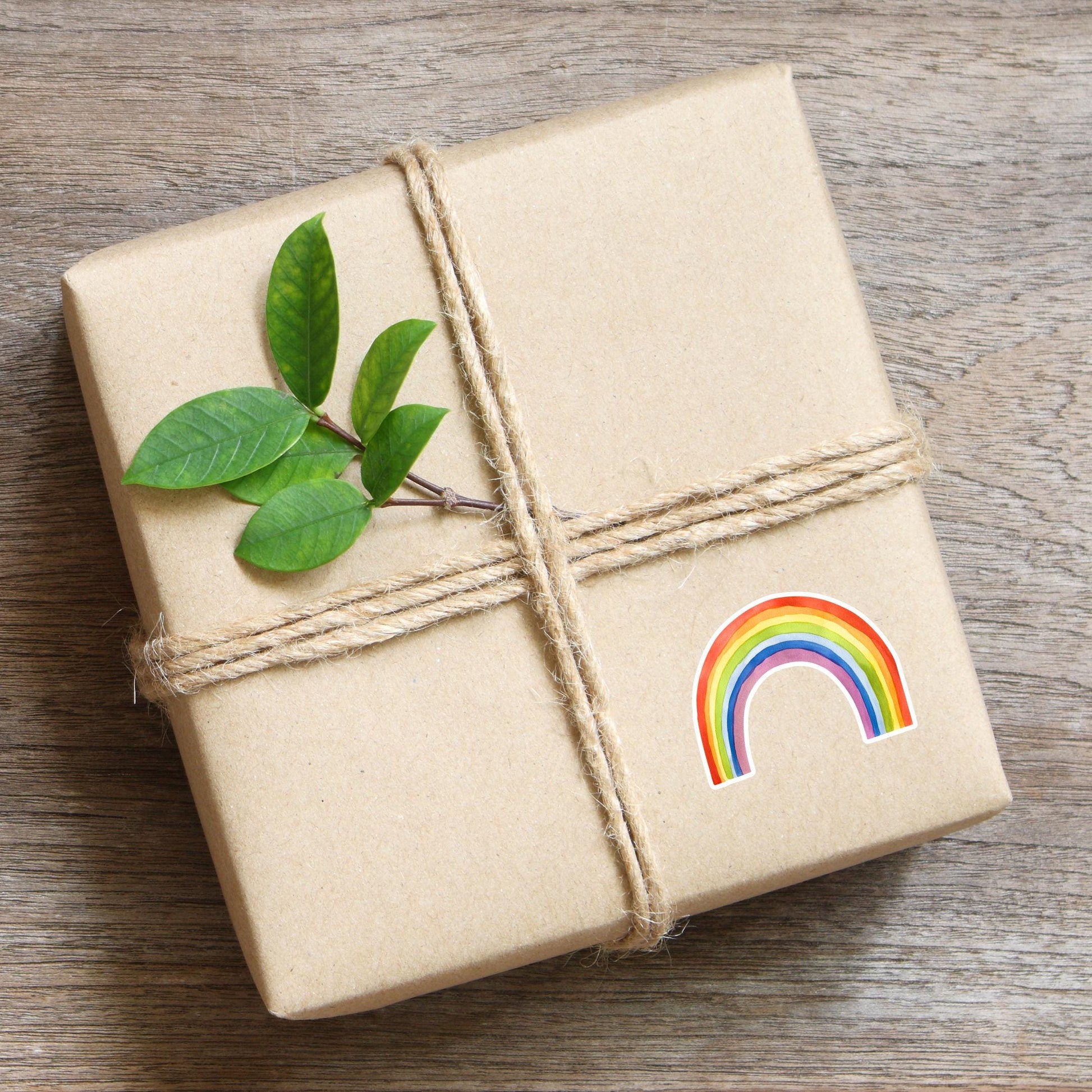 Geschenk in Kraftpapier mit Juteschnur gebunden, verziert mit einem frischen Zweig und einem Sticker Regenbogen, handgemalt, wasserfest, ideal für kreative Dekorationen.