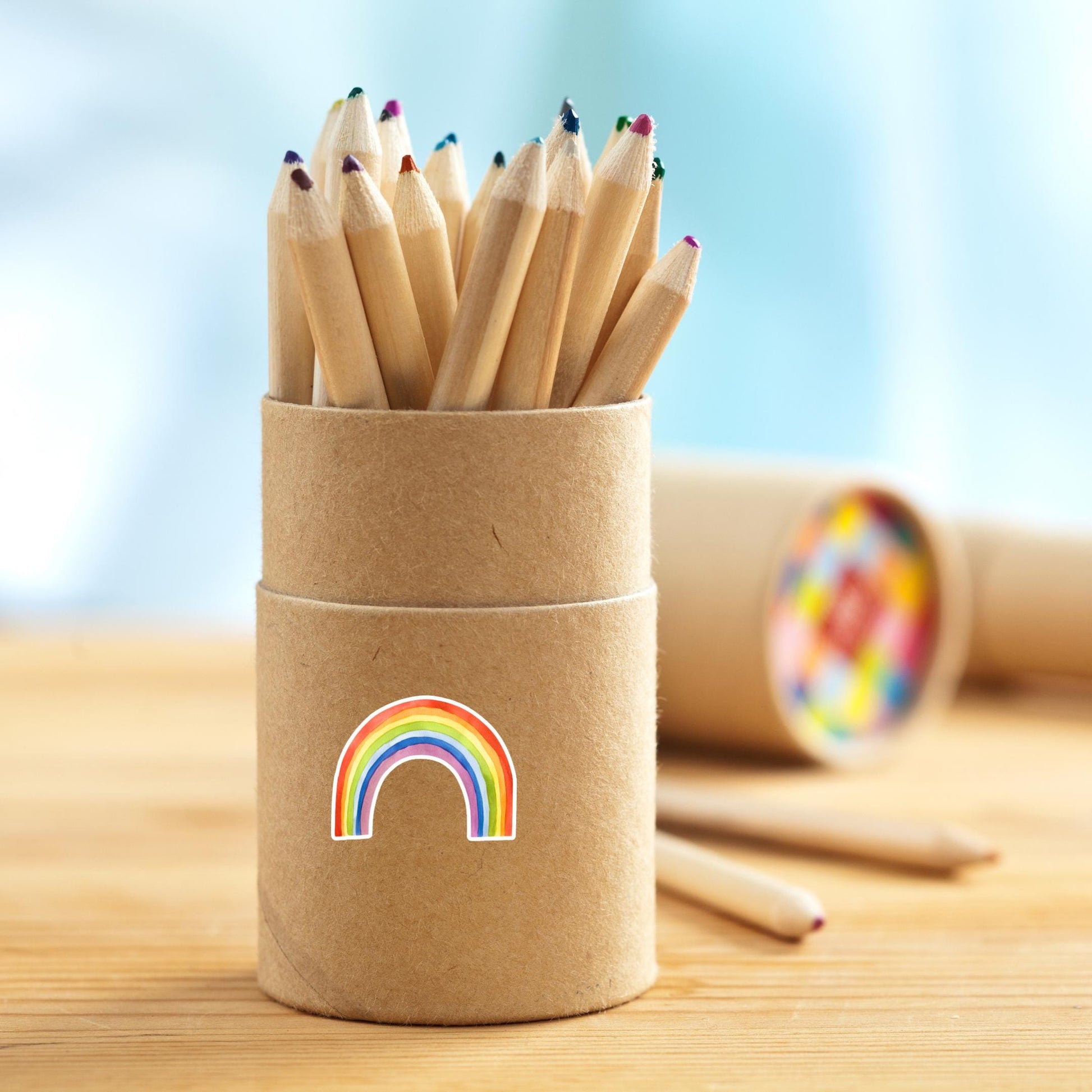 Runde Stifteköcher aus Kraftpapier mit naturfarbenen Buntstiften, dekoriert mit einem Sticker Regenbogen. Zwei lose Stifte liegen daneben.