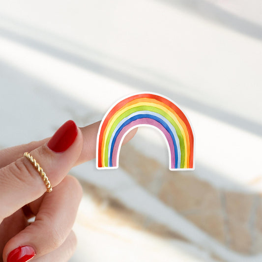 Hand hält Sticker Regenbogen mit sechs Farbbändern. Roter Nagellack und goldener Ring sichtbar. Handgemalt, langlebig, ideal zum Dekorieren von Notizbüchern oder Laptops.