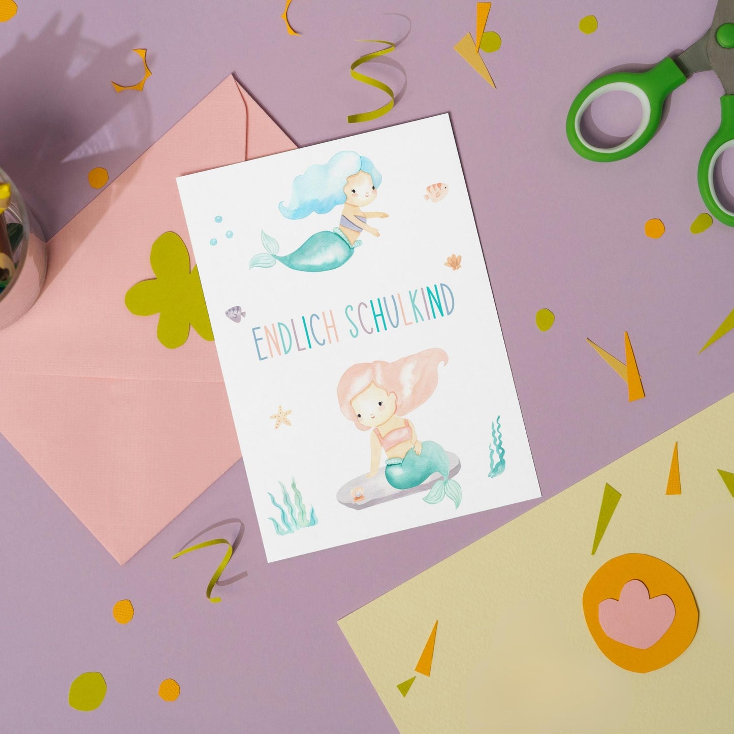 Karte Endlich Schulkind Meerjungfrau A6: Pastellfarbene Postkarte mit Meerjungfrauen-Design und Schriftzug, umgeben von Bastelmaterial, inklusive passendem rosa Umschlag und grüner Schere.
