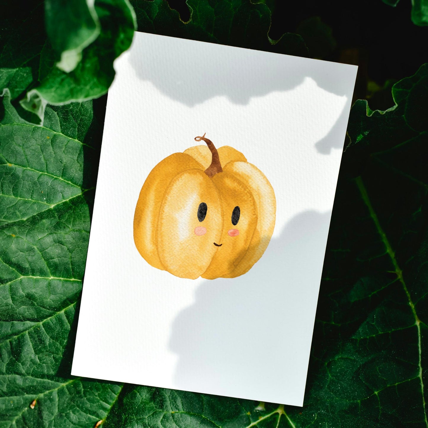 Postkarten Set Kürbis A6 zeigt eine Karte mit einem lächelnden Kürbis auf weißem Papier, platziert auf dunklen Blättern. Ideal für Herbstgrüße.