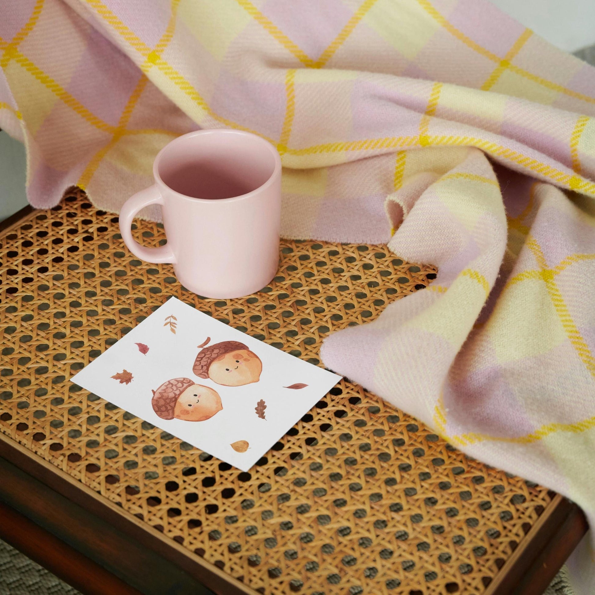 Karte Eicheln & Herbstlaub A6 auf kleinem, geflochtenem Tablett, begleitet von einer rosa Keramiktasse und einer pastellfarbenen Decke im Hintergrund.