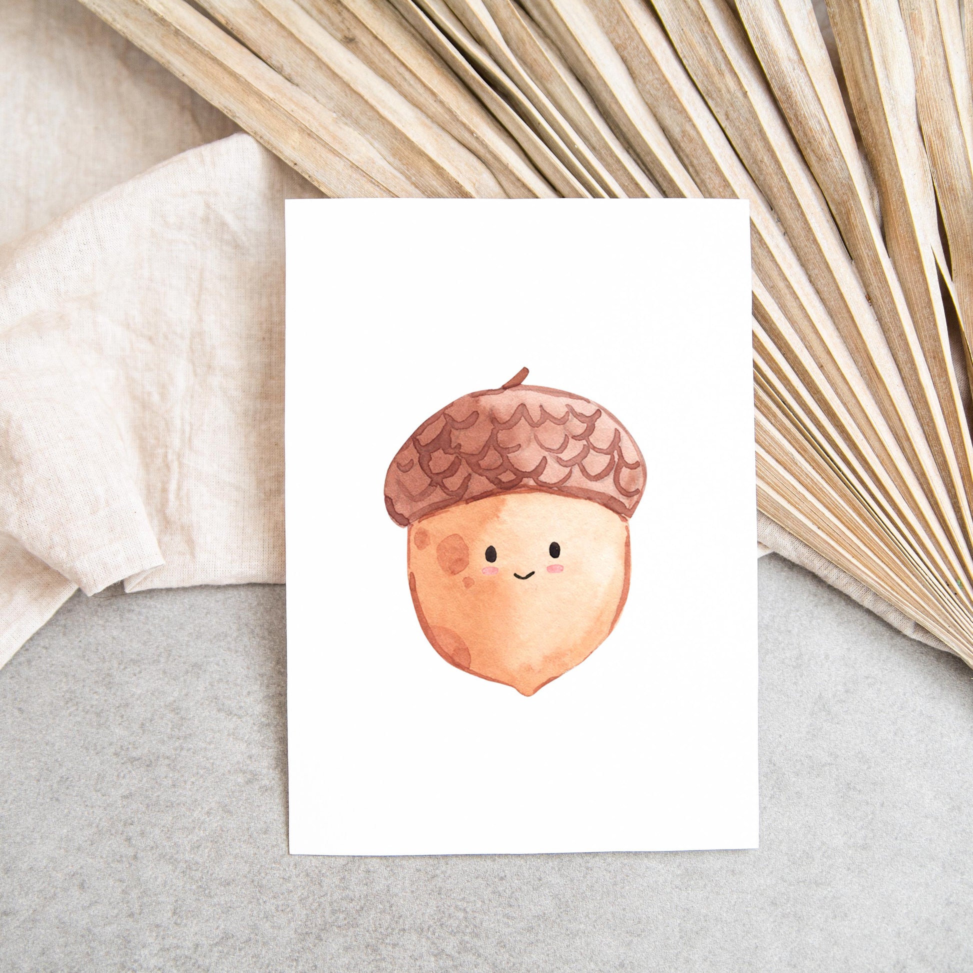 Karte Eichel A6 zeigt eine handgemalte, lächelnde Eichel auf weißem Papier, umgeben von Leinen und getrockneten Palmwedeln. Perfekt als Glückwunschkarte oder Herbstdeko.