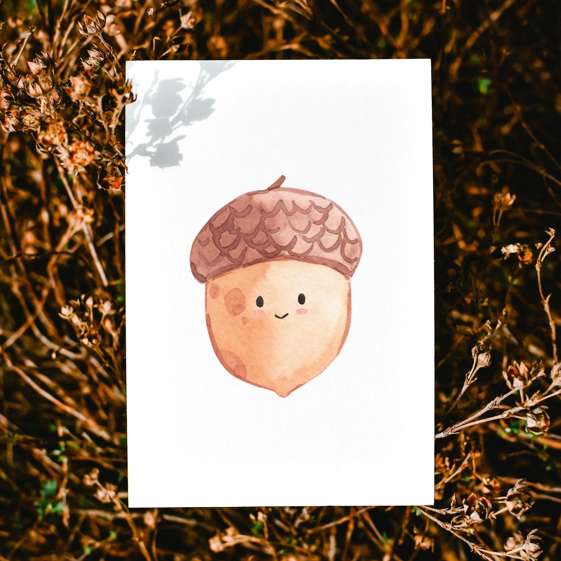 Karte Eichel A6 auf getrocknetem Gras, zeigt eine lächelnde Aquarell-Eichel mit Gesicht, gedruckt auf hochwertigem 300g Naturpapier.