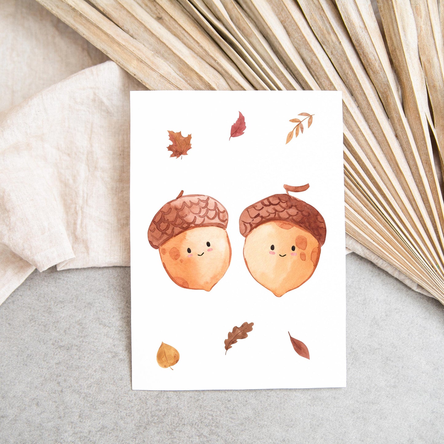 Karte Eicheln & Herbstlaub A6 zeigt zwei lächelnde Aquarell-Eicheln mit Herbstblättern auf beigem Stoff, ideal für Grüße oder Dekoration.