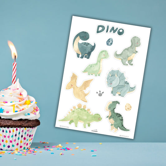 Ausdruck von gratis Cake Topper zum kostenlosen Download mit niedlichen Aquarell Dinosauriern liegt neben einem Cupcake mit weißem Zuckerguss, Streuseln und einer Geburtstagskerze.