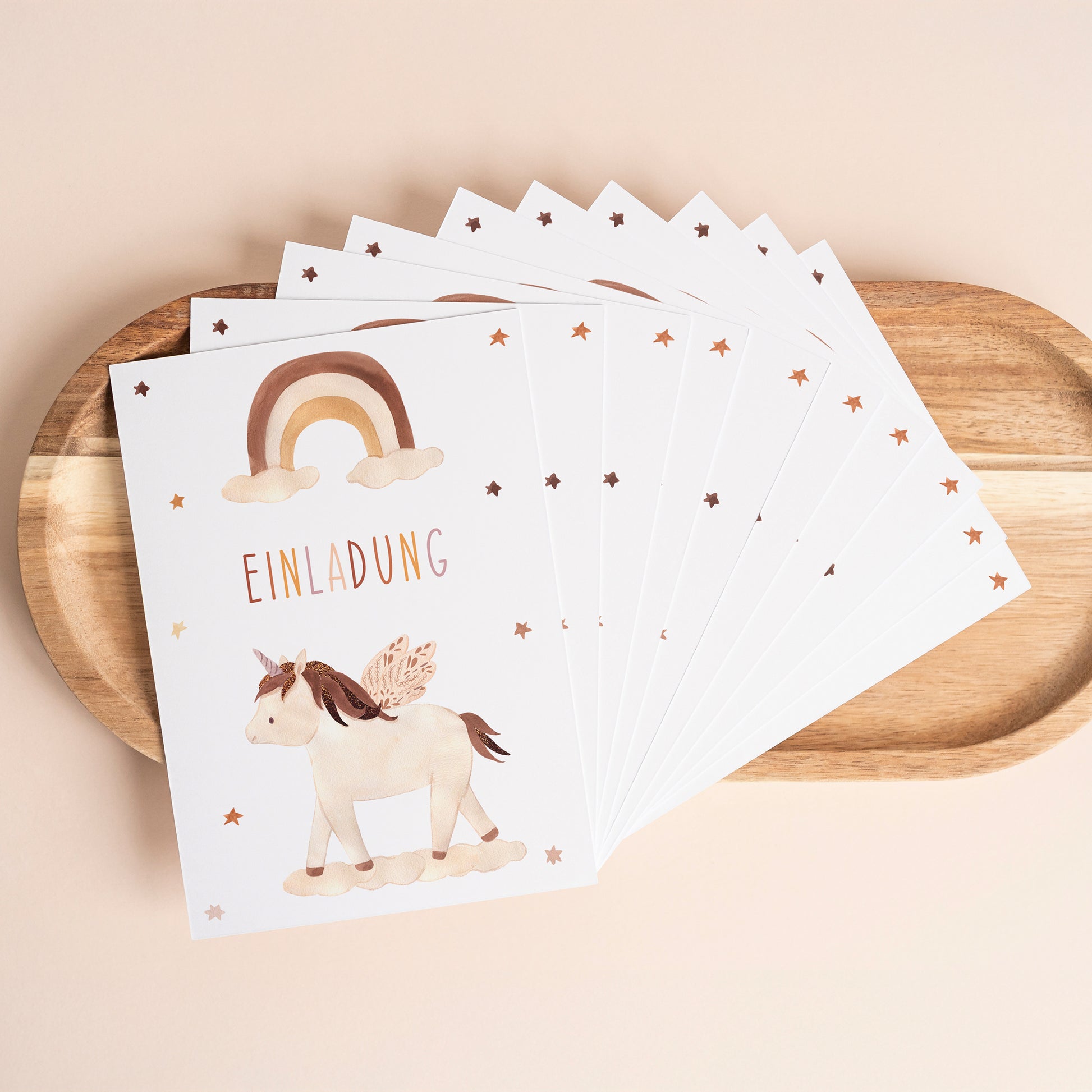 Einladung Kindergeburtstag Einhorn Boho A6: Holztablett mit weißen Einladungskarten, pastellfarbenem Regenbogen, Sternen und einem geflügelten Einhorn auf Wolke.