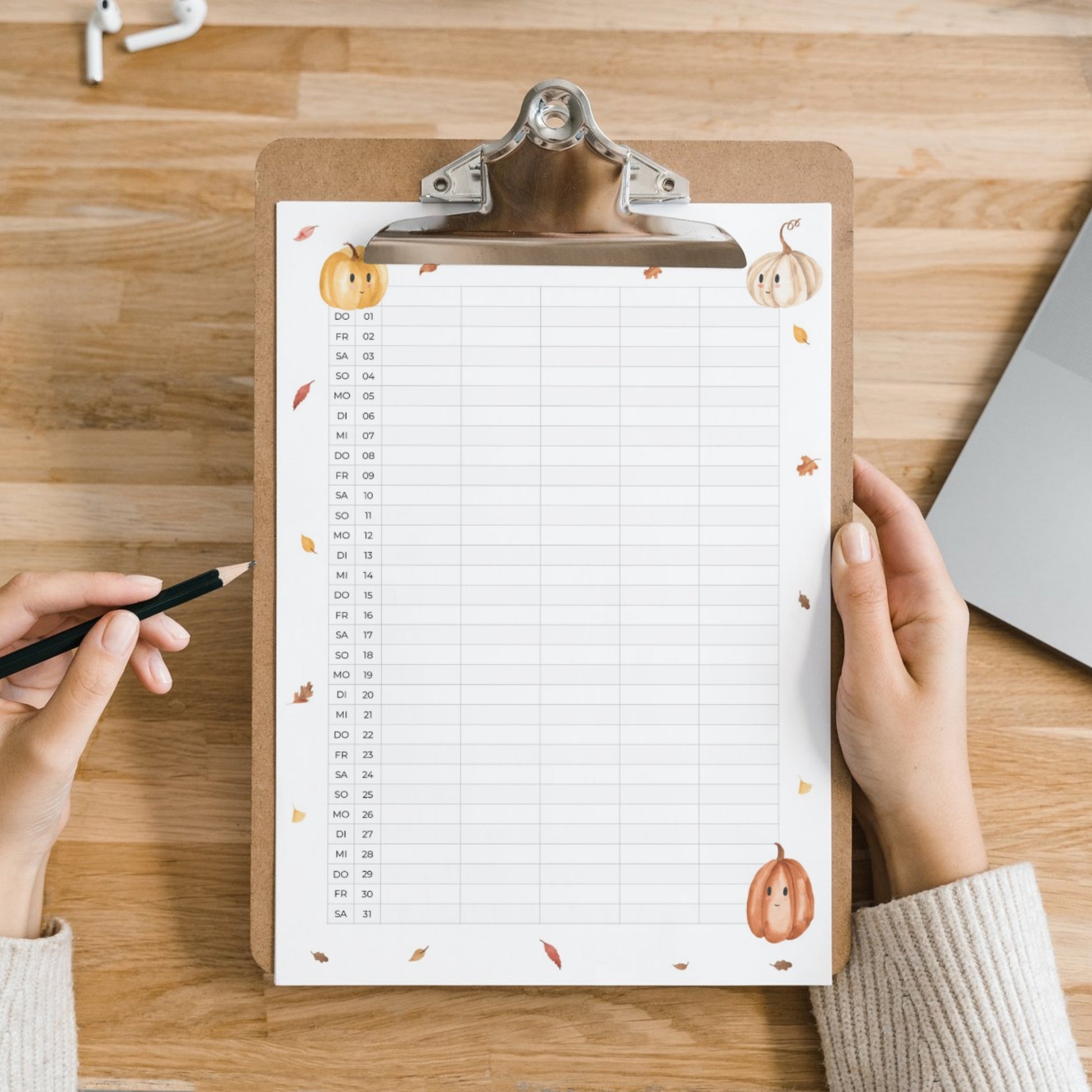 Familienkalender 2026 zum Ausdrucken 5 Spalten auf Klemmbrett mit Aquarellkürbissen, daneben liegen kabellose Ohrhörer, Laptop-Ecke sichtbar. Hände halten Klemmbrett und Stift.