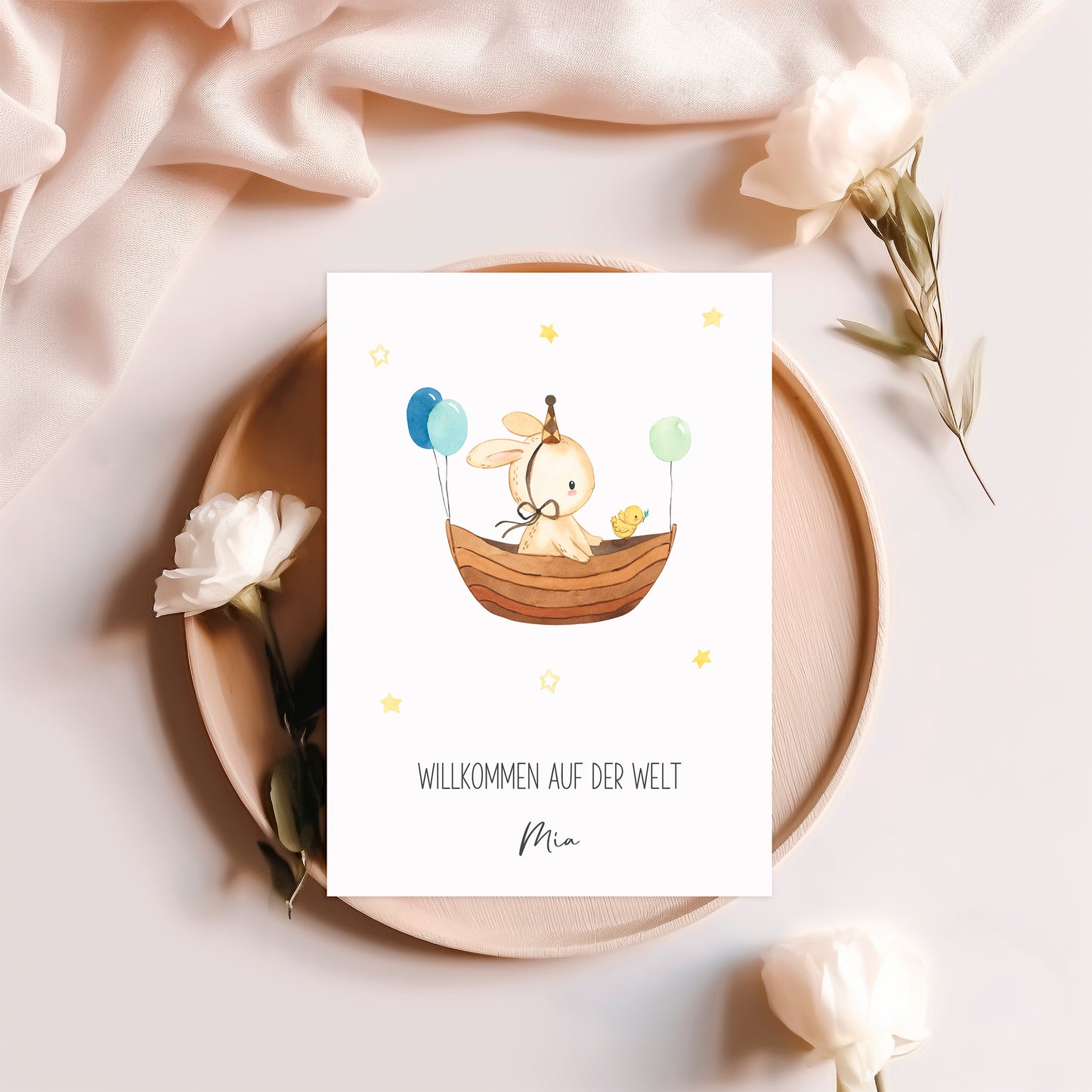 Personalisierte Geburtskarte Hase & Luftschiff A6, zeigt einen Hasen im Boot mit Küken und Ballons, umgeben von Blumen auf einem Holzteller.