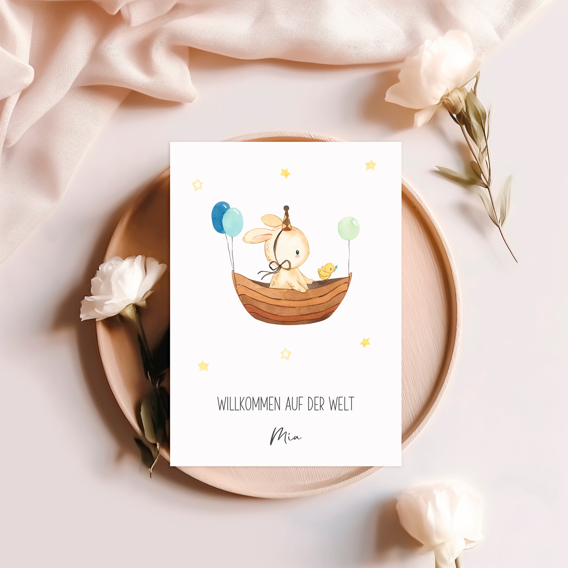 Personalisierte Geburtskarte Hase & Luftschiff A6, zeigt einen Hasen im Boot mit Küken und Ballons, umgeben von Blumen auf einem Holzteller.