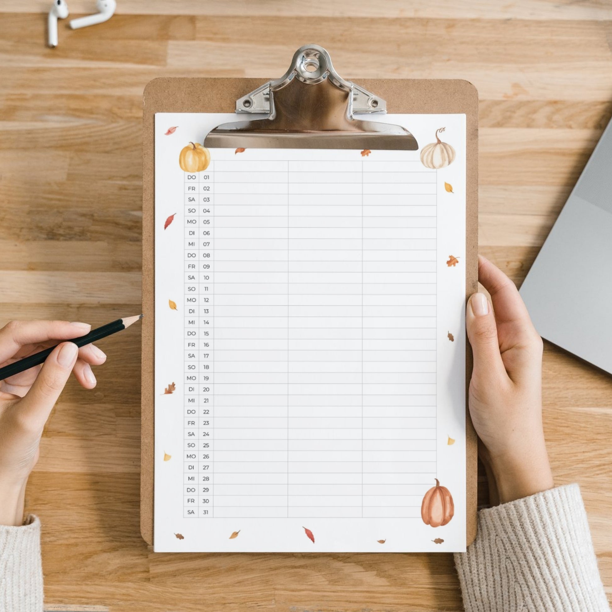 Familienkalender 2026 zum Ausdrucken 3 Spalten auf einem Holzschreibtisch, dekoriert mit Herbstillustrationen, gehalten von Händen mit Stift, neben Laptop und Ohrstöpseln.
