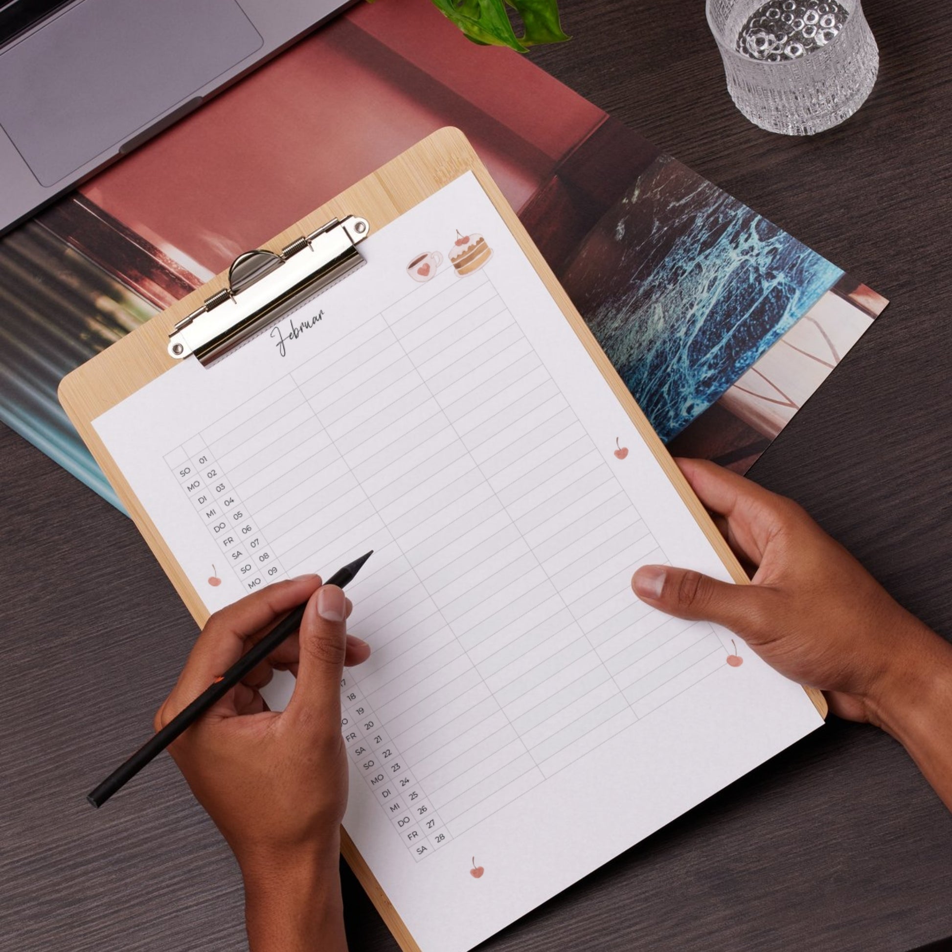 Hände halten ein Klemmbrett mit Paarkalender 2026 zum Ausdrucken A3 & A4, Februarblatt, umgeben von Laptop, Fotos, Pflanze und Glas.