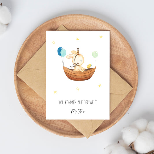 Personalisierte Geburtskarte Hase & Luftschiff A6 auf Holzplatte, zeigt Hase im Boot mit Ballons, neben Küken, mit WILLKOMMEN AUF DER WELT Matteo Schriftzug.