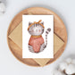 Karte Katze Vintage A6: Handgezeichnete Karte mit einer Katze in einem Pullover und mit Schleife, auf einer Holzplatte präsentiert. Ideal als Gruß- oder Geburtstagskarte.