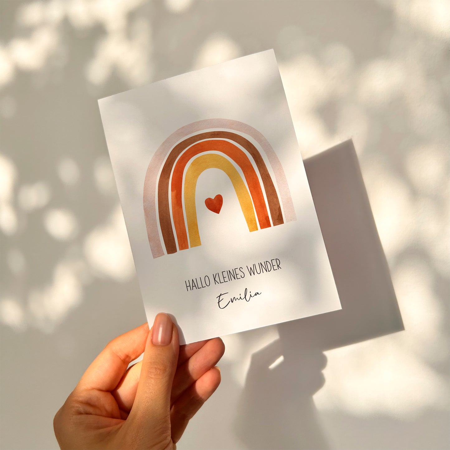 Personalisierte Geburtskarte Regenbogen & Herz A6 zeigt handgehaltene Karte mit Regenbogen, Herz und Hallo kleines Wunder in Deutsch, Name Emilia darunter.