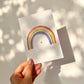 Hand hält Karte Regenbogen mit Herz A6, handbemalt auf hochwertigem, mattem Naturpapier. Perfekt für Grüße oder Glückwünsche. Größe A6, Rückseite beschreibbar.