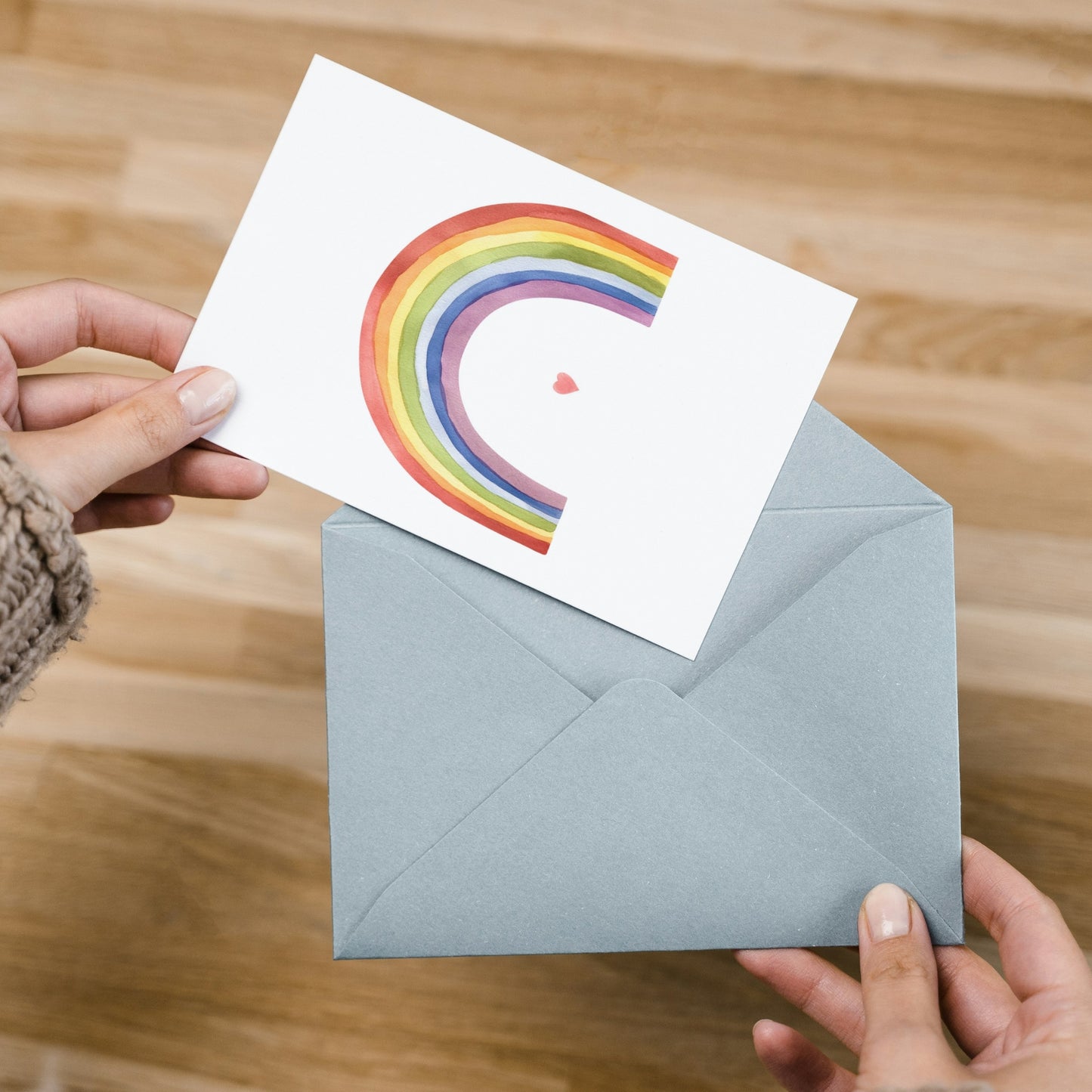 Hände ziehen die Karte Regenbogen mit Herz A6 aus einem grauen Umschlag, zeigt handgezeichneten Regenbogen und Herz, auf Holztisch liegend.