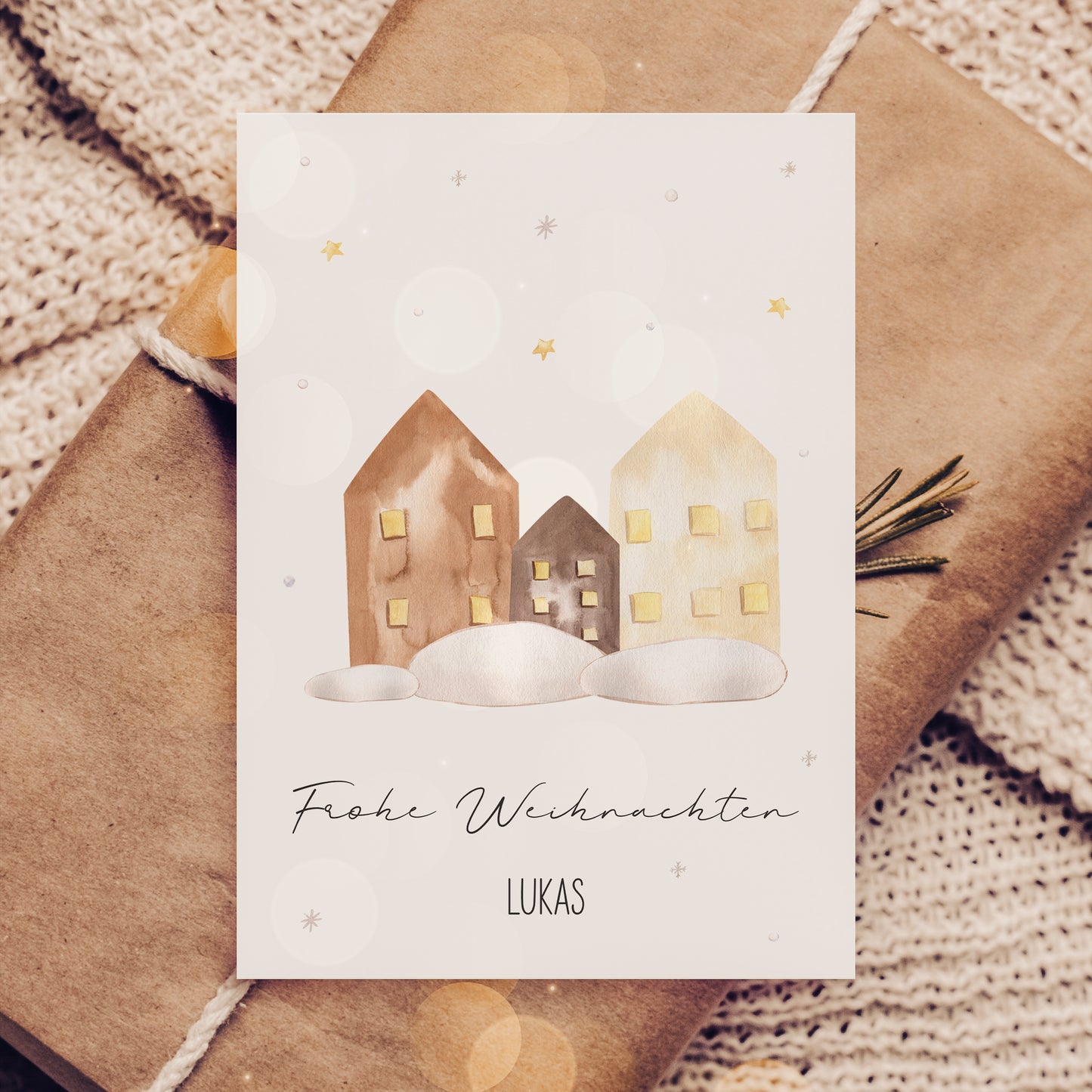 Personalisierte Weihnachtskarte Häuser A6 auf braunem Kraftpapier, zeigt drei Aquarellhäuser im Schnee mit goldenem Sternenhimmel und Frohe Weihnachten, Lukas in kursiver Schrift.