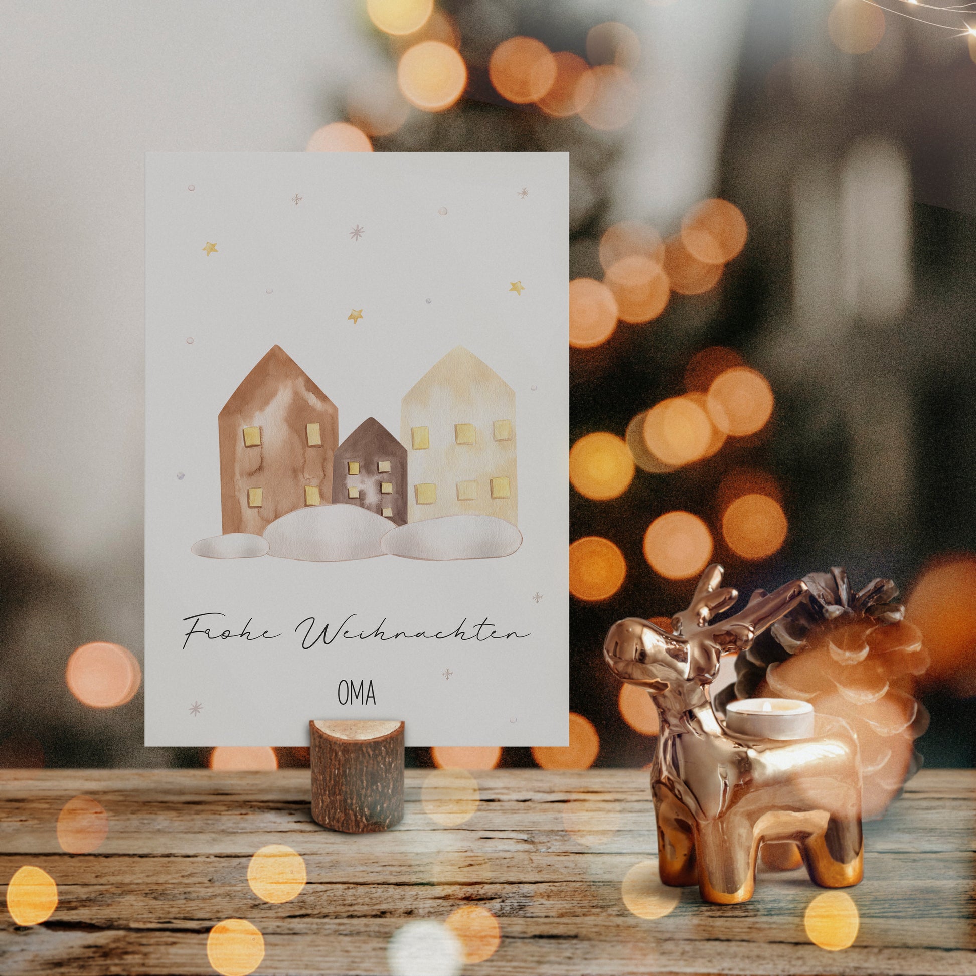 Weihnachtskarte Personalisierte Weihnachtskarte Häuser A6 auf Holzständer, mit Aquarellhäusern und Frohe Weihnachten OMA, neben einem Rentier-Kerzenhalter und Tannenzapfen.