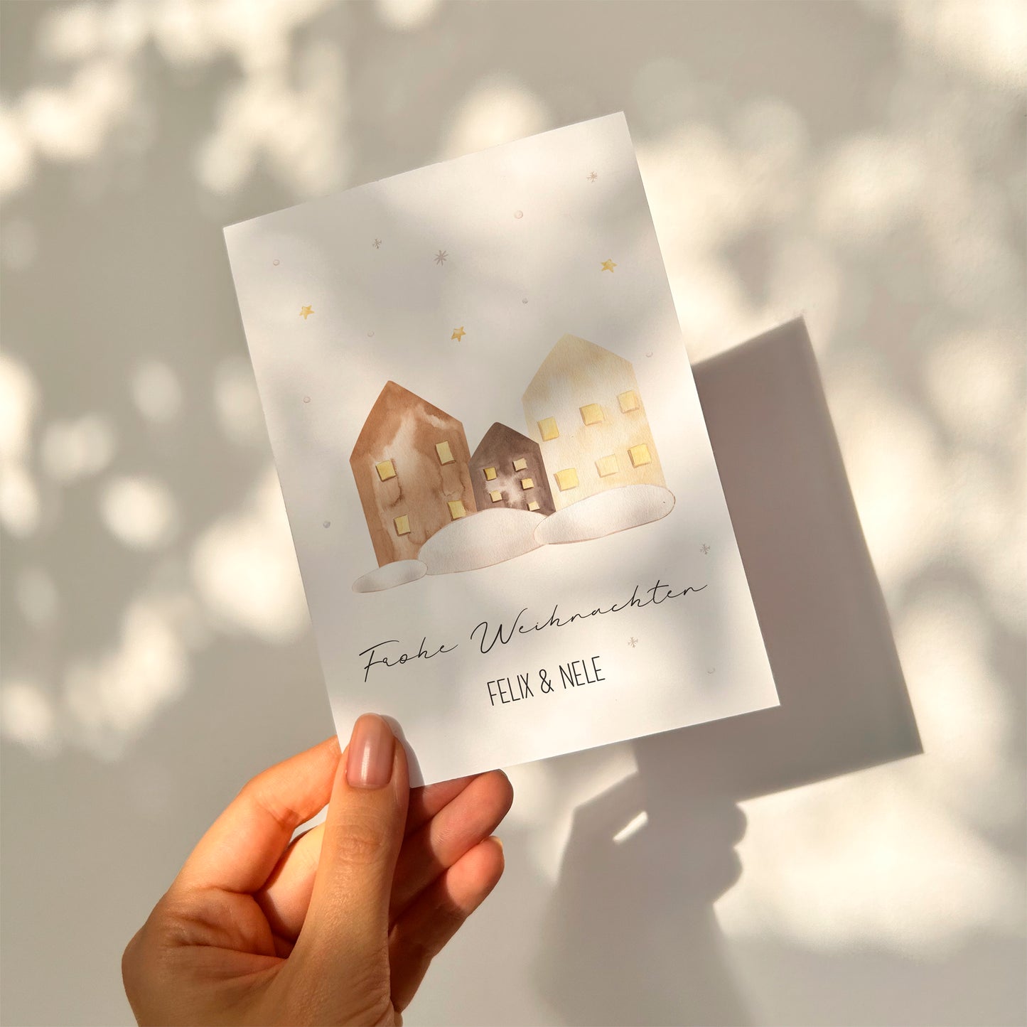 Personalisierte Weihnachtskarte Häuser A6; zeigt handgemalte Häuser im Schnee mit Frohe Weihnachten und Namen, gehalten von einer Hand vor einer sanft beleuchteten Wand.