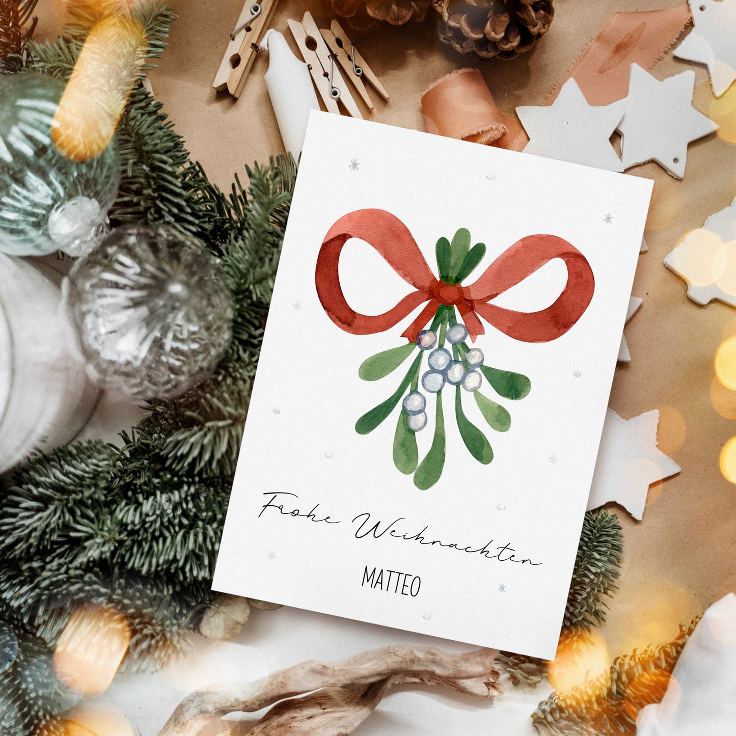 Personalisierte Weihnachtskarte Mistelzweig A6 zeigt handgemalte Mistelzweig mit roter Schleife, umgeben von weihnachtlichen Dekorationen auf Bastelpapiert.