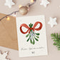 Personalisierte Weihnachtskarte Mistelzweig A6 mit Aquarell-Motiv, beigelegtem Kraftpapier-Umschlag, umgeben von weihnachtlichen Dekorationen wie Bauble, Lichtern und Tannenzweigen.